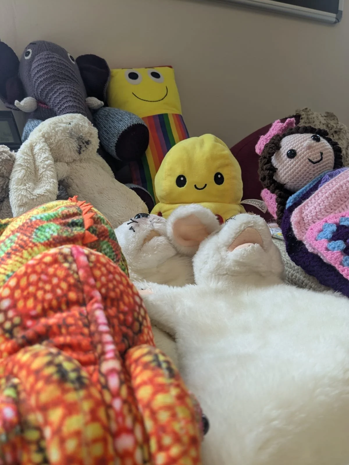 A collection of plush toys and stuffed animals arranged on a bed or surface, including colorful animals, a smiley face pillow, and a white fluffy plush with its paws up.
