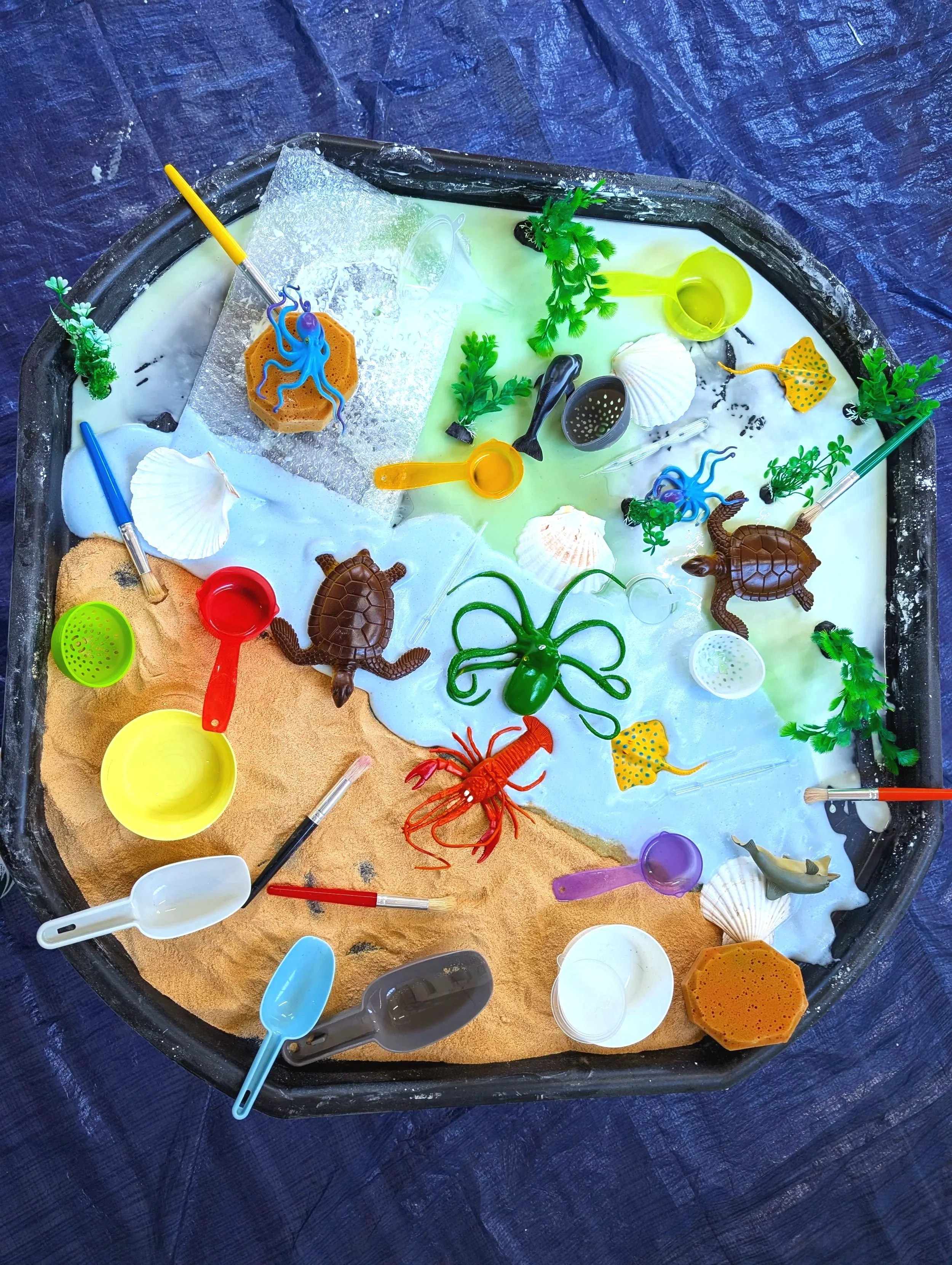 A mixed underwater-themed sandbox with plastic sea life toys, sand, and water, including shells, turtles, octopuses, fish, and small buckets and shovels.