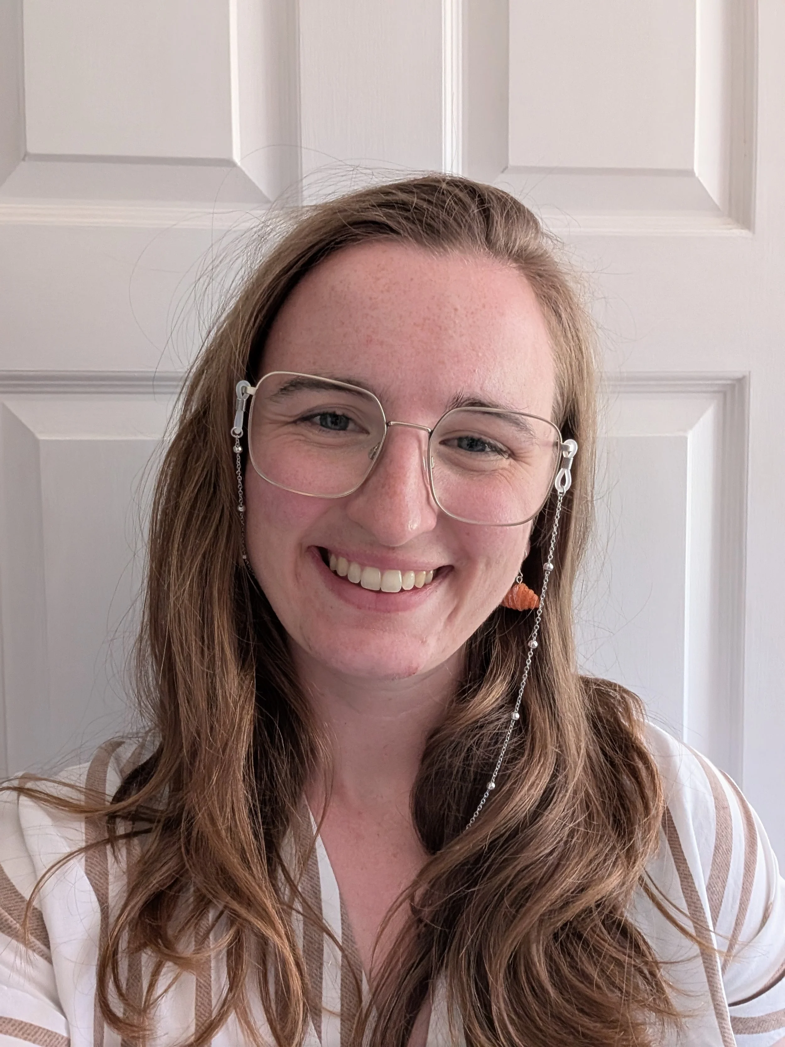 A woman with long, wavy hair and glasses smiling in front of a white door.