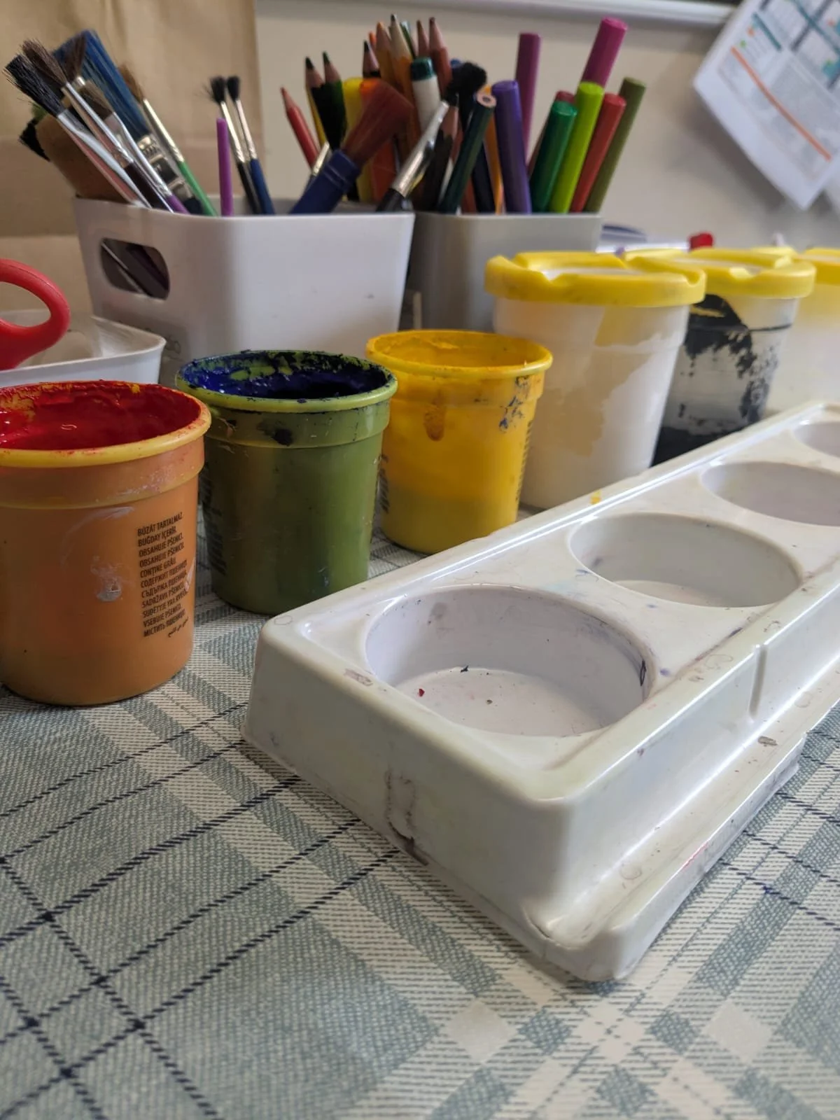 Paint palette with empty paint wells, jars of colorful paint, and paintbrushes in containers on a checkered tablecloth.