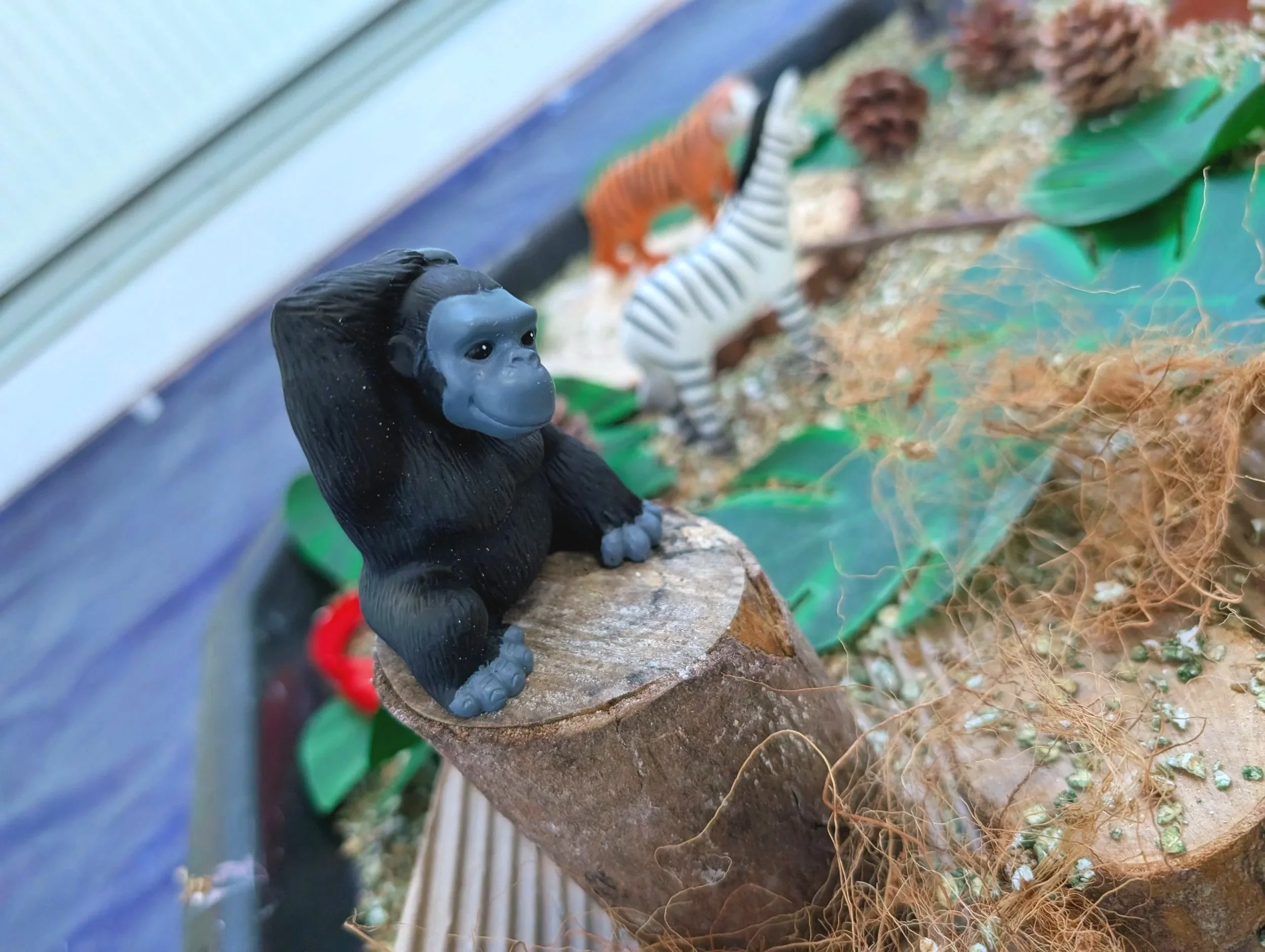 Close-up of a black monkey figurine with a light blue face, sitting on a wooden surface with a jungle-themed background including other animal figures, plants, and natural elements.