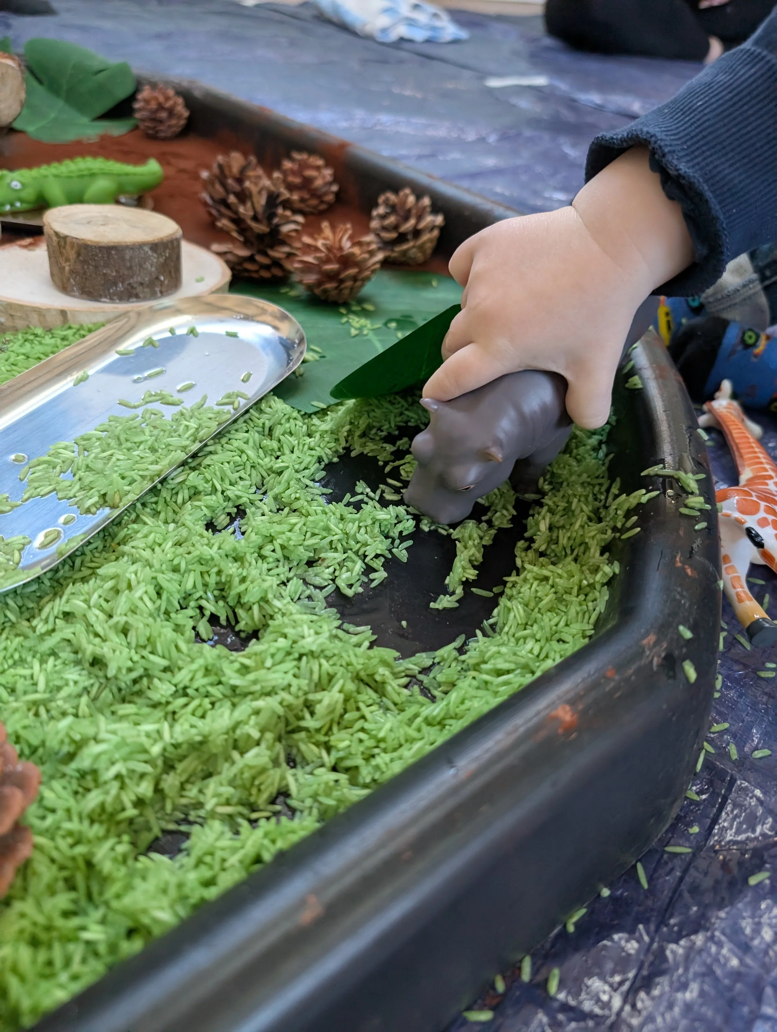 A child's hand using a gray plastic hippo to play with green rice-shaped pasta on a large black tray, surrounded by pinecones, green leaves, and small toy animals.