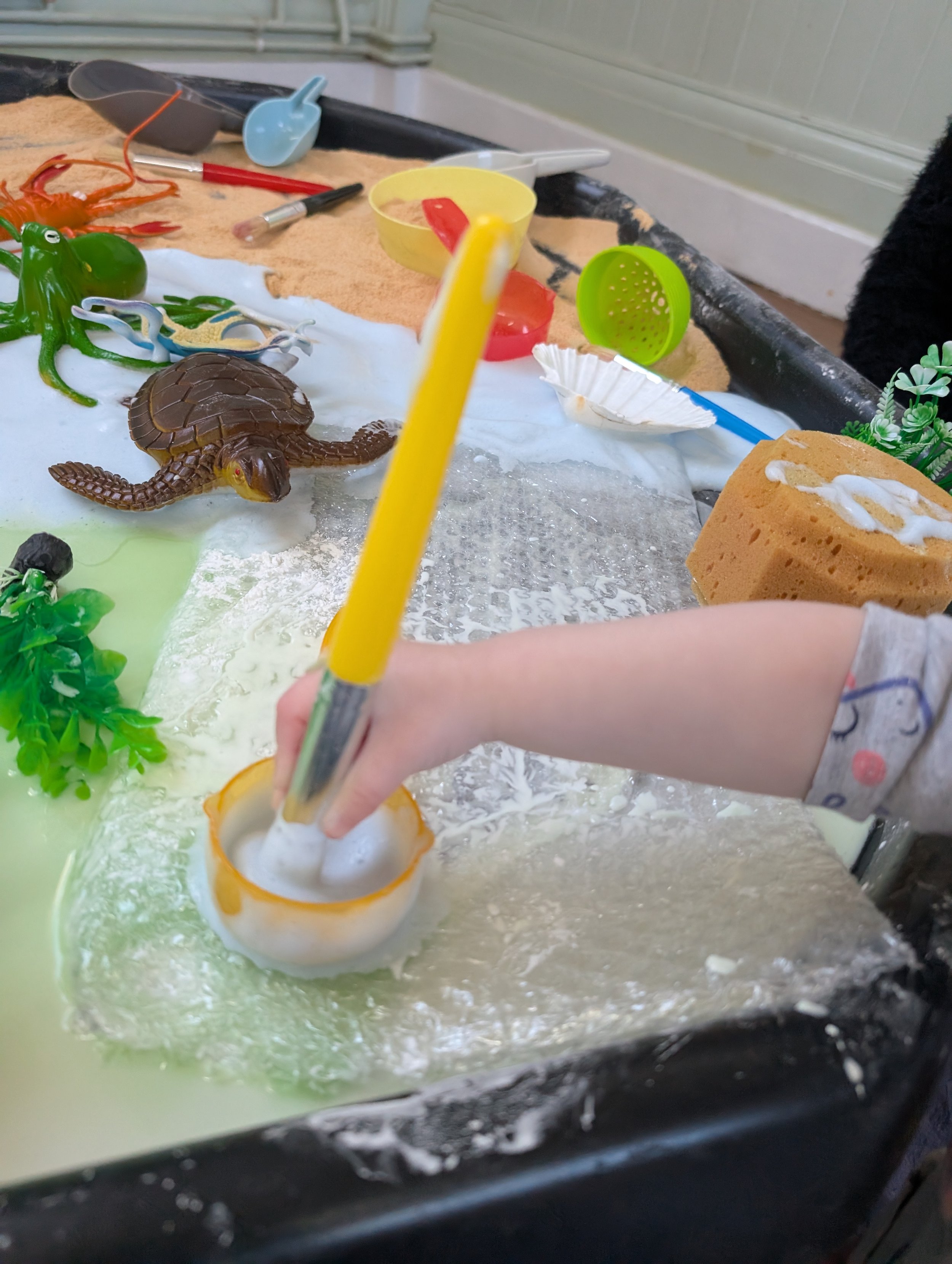Close-up of a children's sandbox with toy animals, plastic shovels, and watering cans. A child's hand with a yellow paintbrush is visible. The sandbox surface is covered with gloop, sand and water.