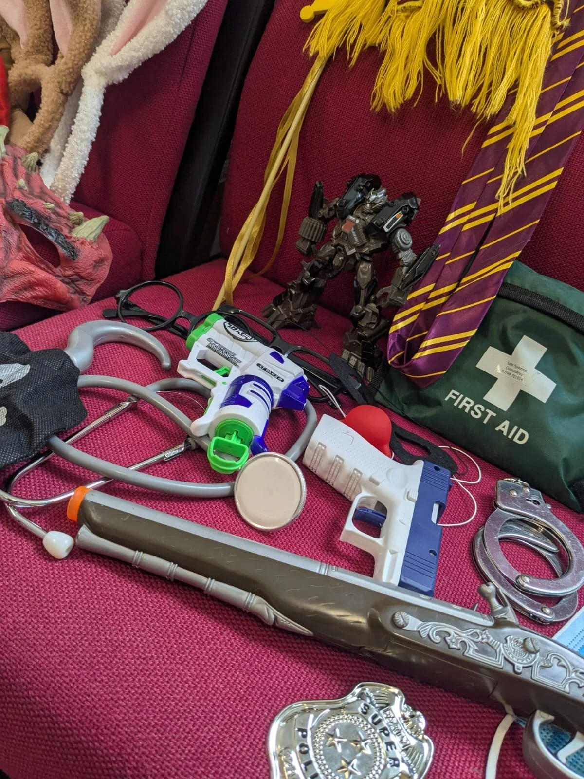 A table covered with various medical and emergency items, including a stethoscope, toy gun, toy robot, first aid kit, handcuffs, and medical tools, on a red fabric surface.