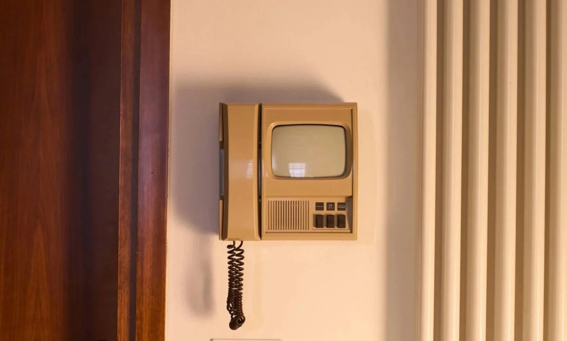 Telefono vintage beige con ricevitore e schermo a tubo catodico, appoggiato su con una parete bianca.