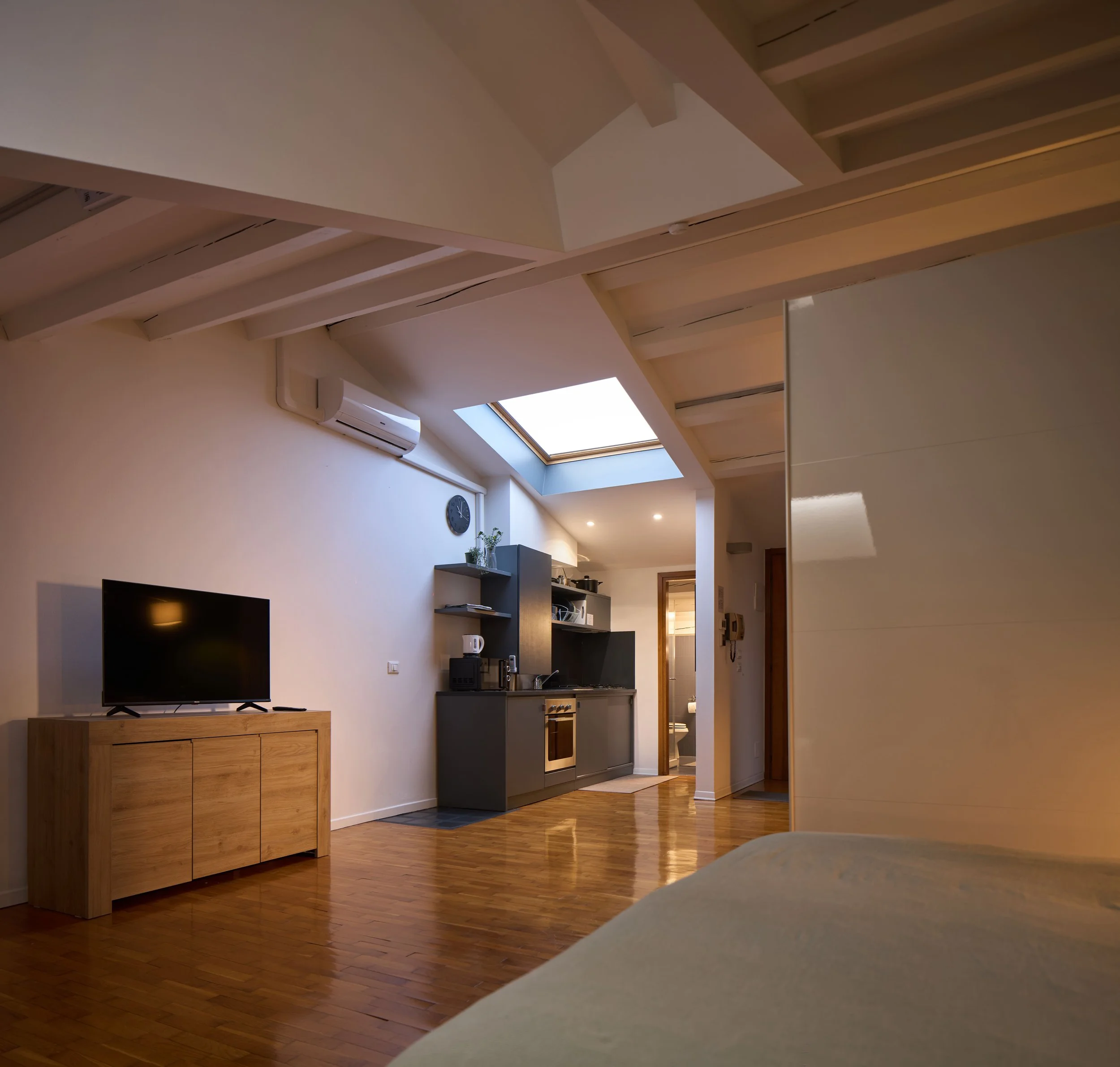 Appartamento con soffitto alto, travi di legno e lucernario, angolo salotto con televisore, mobile in legno, pavimento in legno e cucina visibile sullo sfondo.