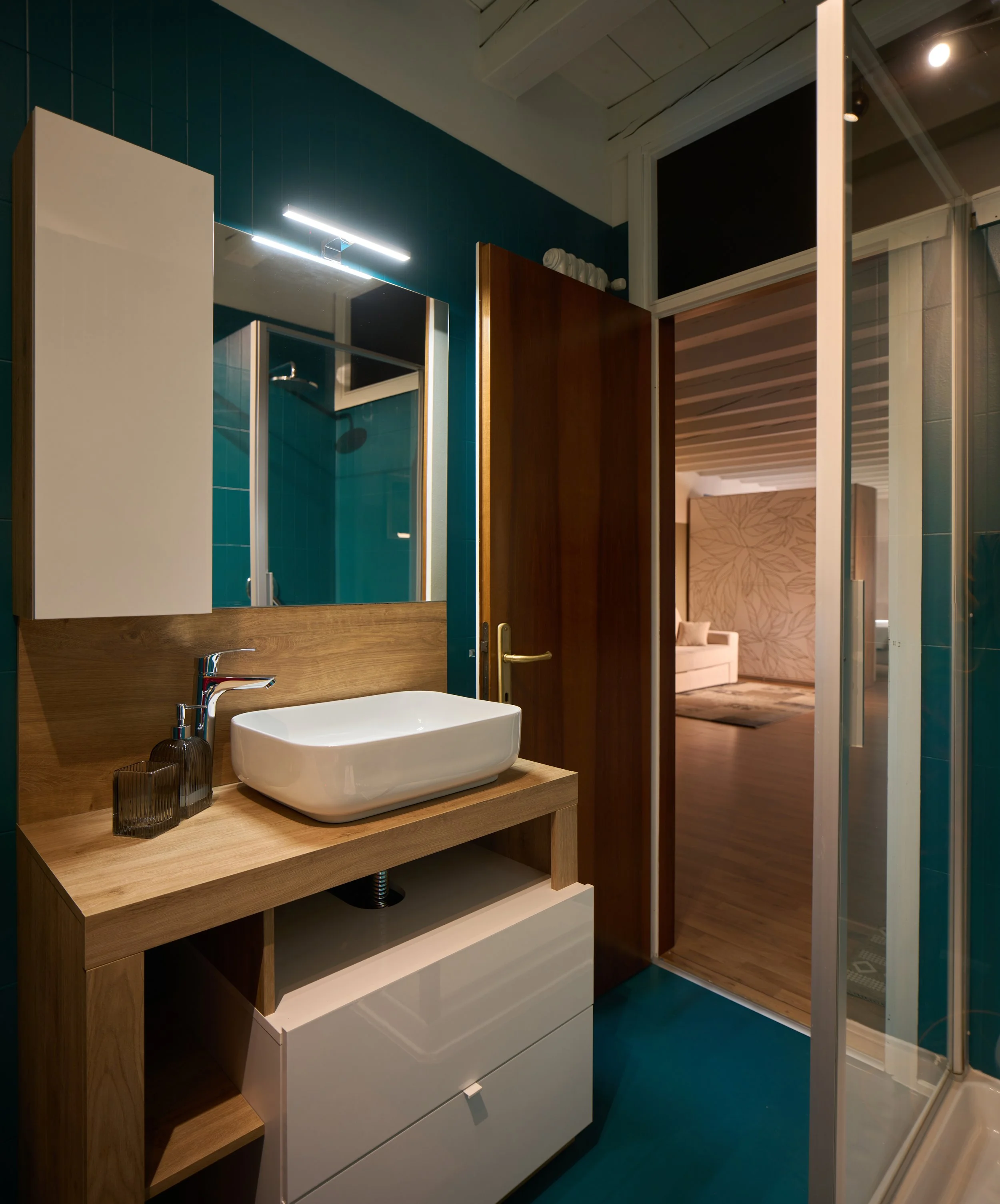 Specchio, lavabo bianco a forma rettangolare su mobile in legno con cassetti bianchi, pareti rivestite in piastrelle blu, porta in legno aperta, vista di un'altra stanza con divano beige e pareti decorate.