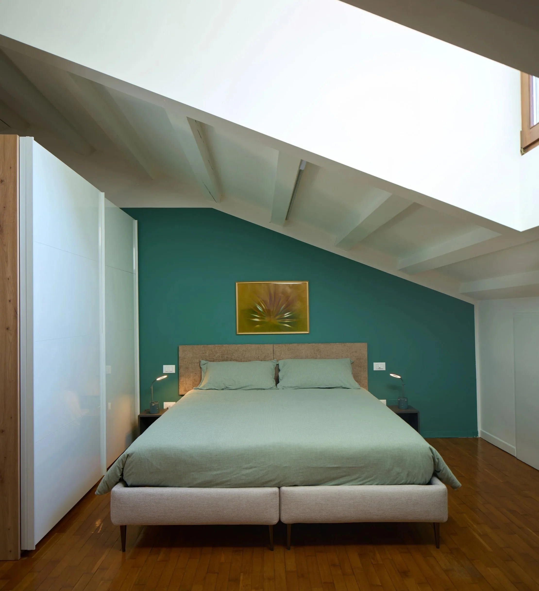 Camera da letto moderna con letto matrimoniale con biancheria verde, due lampade da comodino, pareti verdi, quadro astratto, pavimento in legno, armadio bianco.