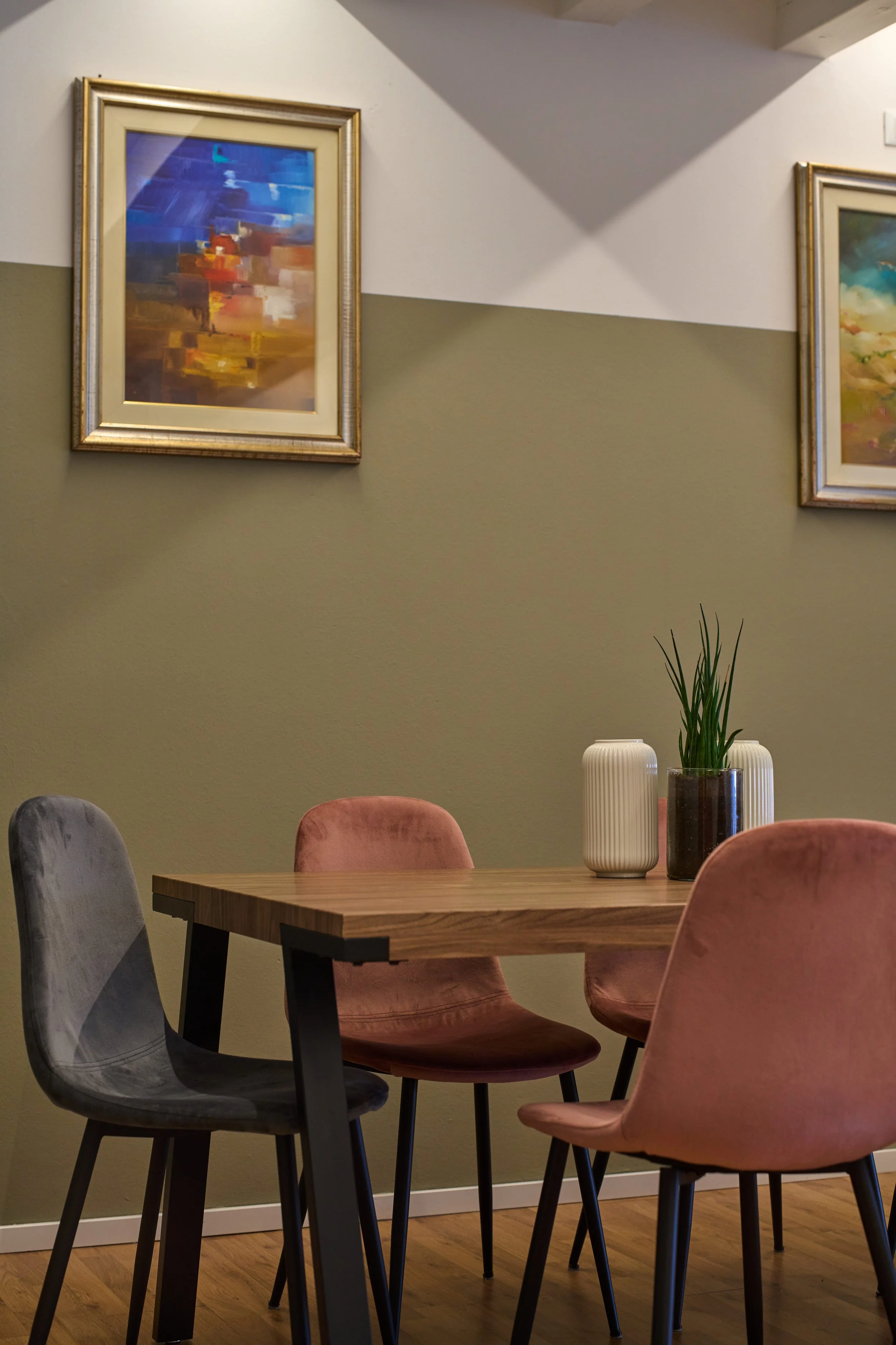 Sala pranzo con quadri astratti appesi e un tavolo con vasi decorativi, sedie di diversi colori e piante.