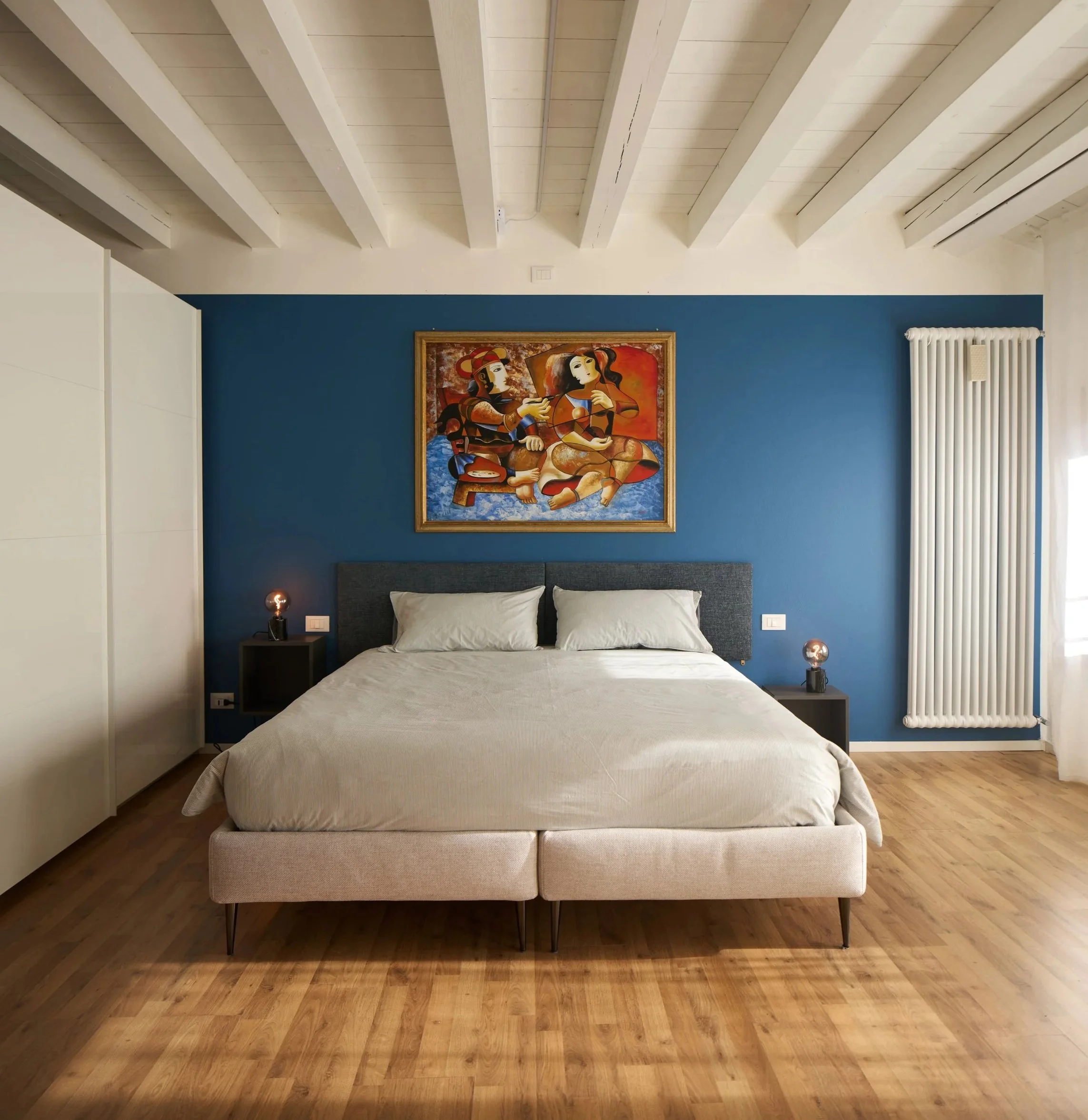 Camera da letto moderna, comodini con lampade, parete blu con quadro colorato, pavimento di legno chiaro.