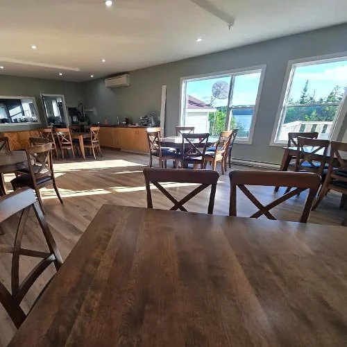 Empty dining area with wooden tables and chairs, large windows with a view of the outside, and sunlight streaming in.