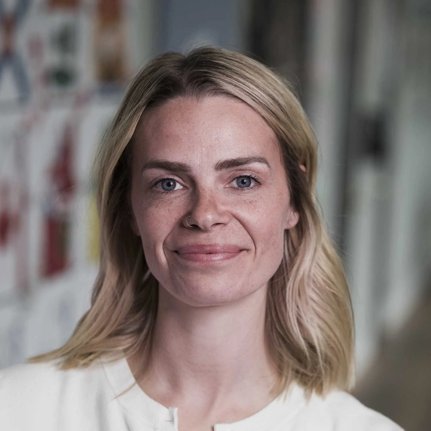 Close-up of a woman with blonde hair, blue eyes, and freckles, smiling at the camera, in an indoor setting with blurred background.