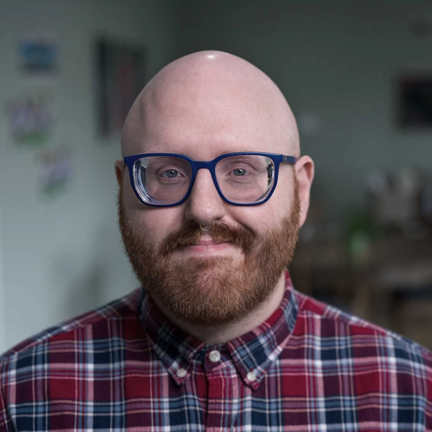 A man with a bald head, glasses, and a beard, wearing a red plaid shirt, smiling indoors.
