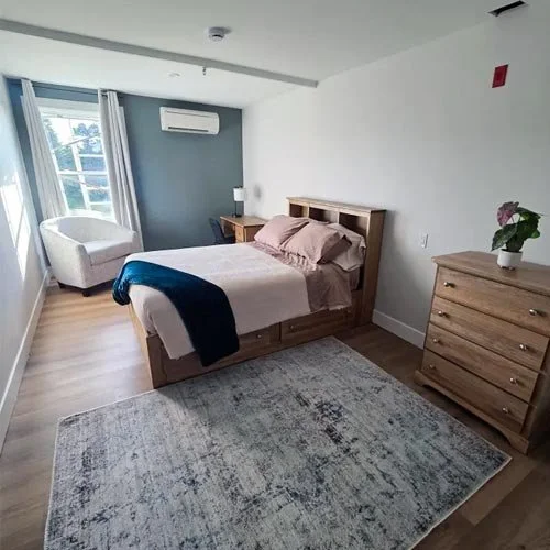 A private bedroom at Vida Nova Recovery Centre. With a bed, wooden dresser, chair, window, air conditioner, and rug.
