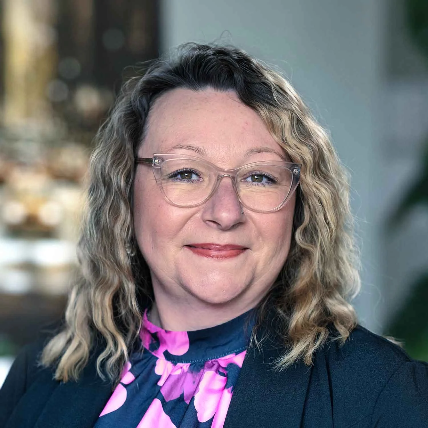 A woman with blond, wavy hair and glasses smiling at the camera, wearing a black jacket and a pink and purple patterned blouse, with a blurred background.