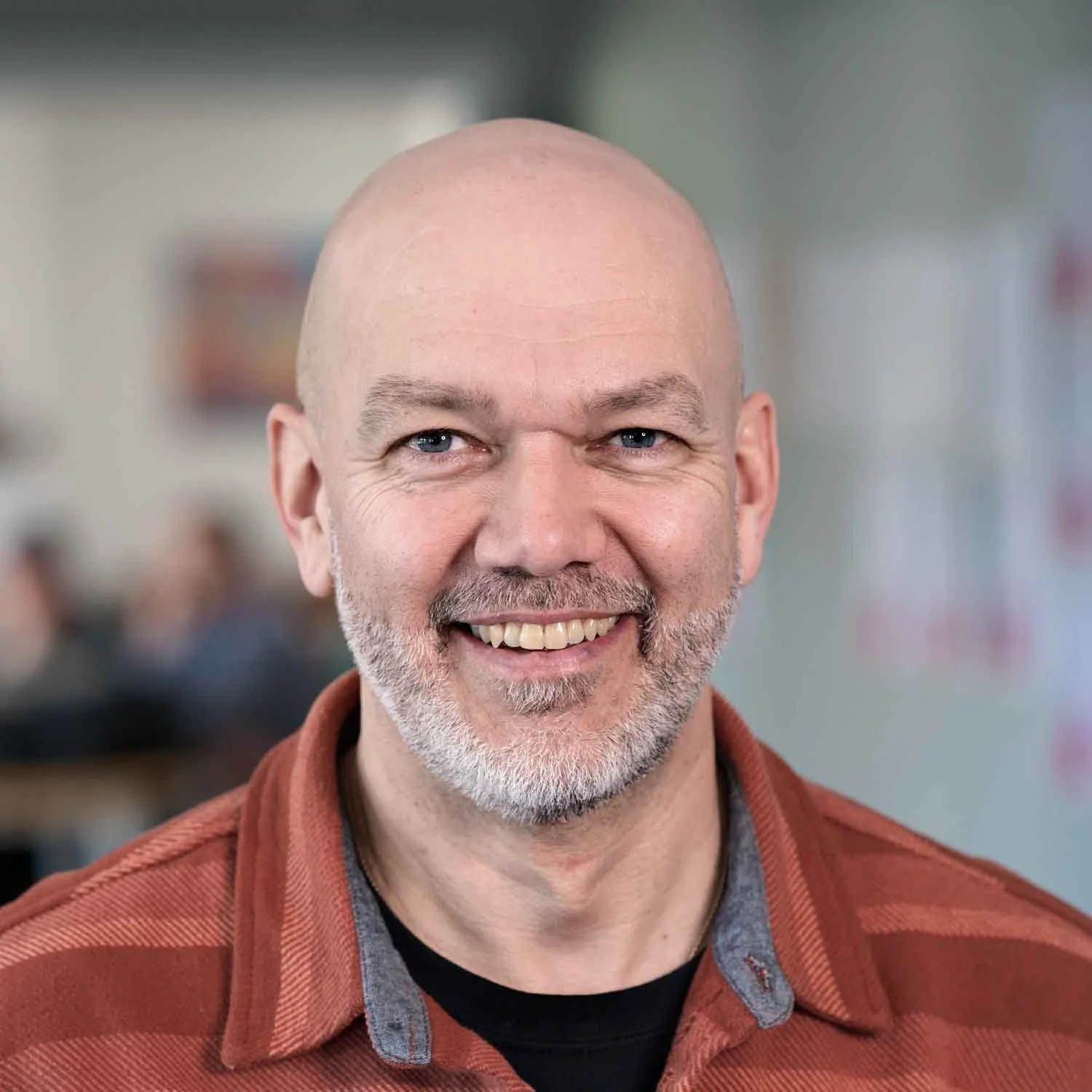 A smiling middle-aged man with a bald head, blue eyes, and a salt-and-pepper beard wearing a rust-colored jacket over a gray shirt, indoors with a blurred background.