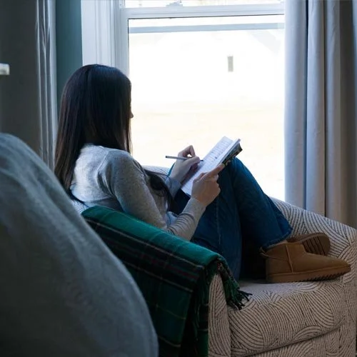A woman sitting on a sofa, reading a book and taking notes, near a large window.