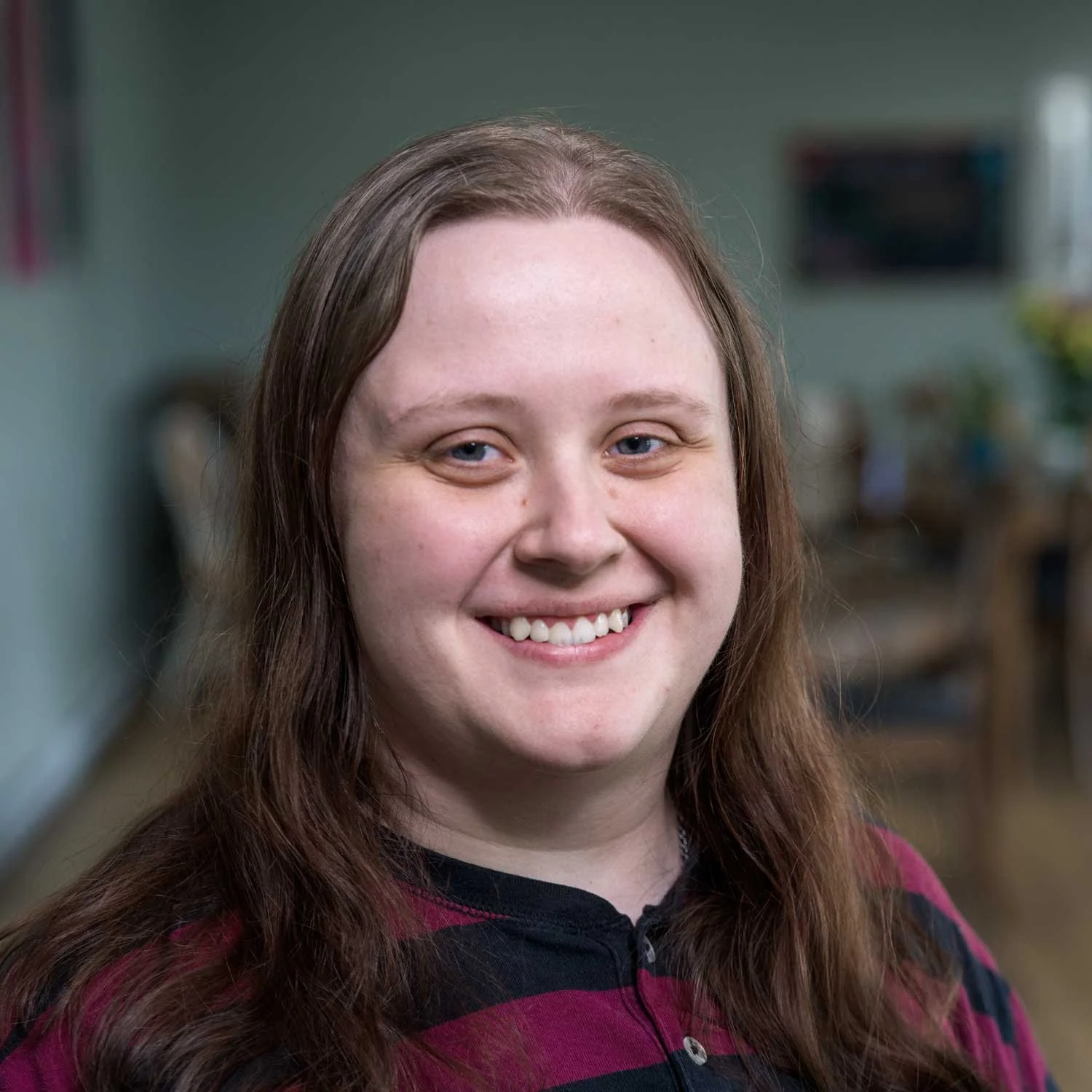 A smiling young woman with long, wavy brown hair and blue eyes, wearing a maroon and black striped hoodie.