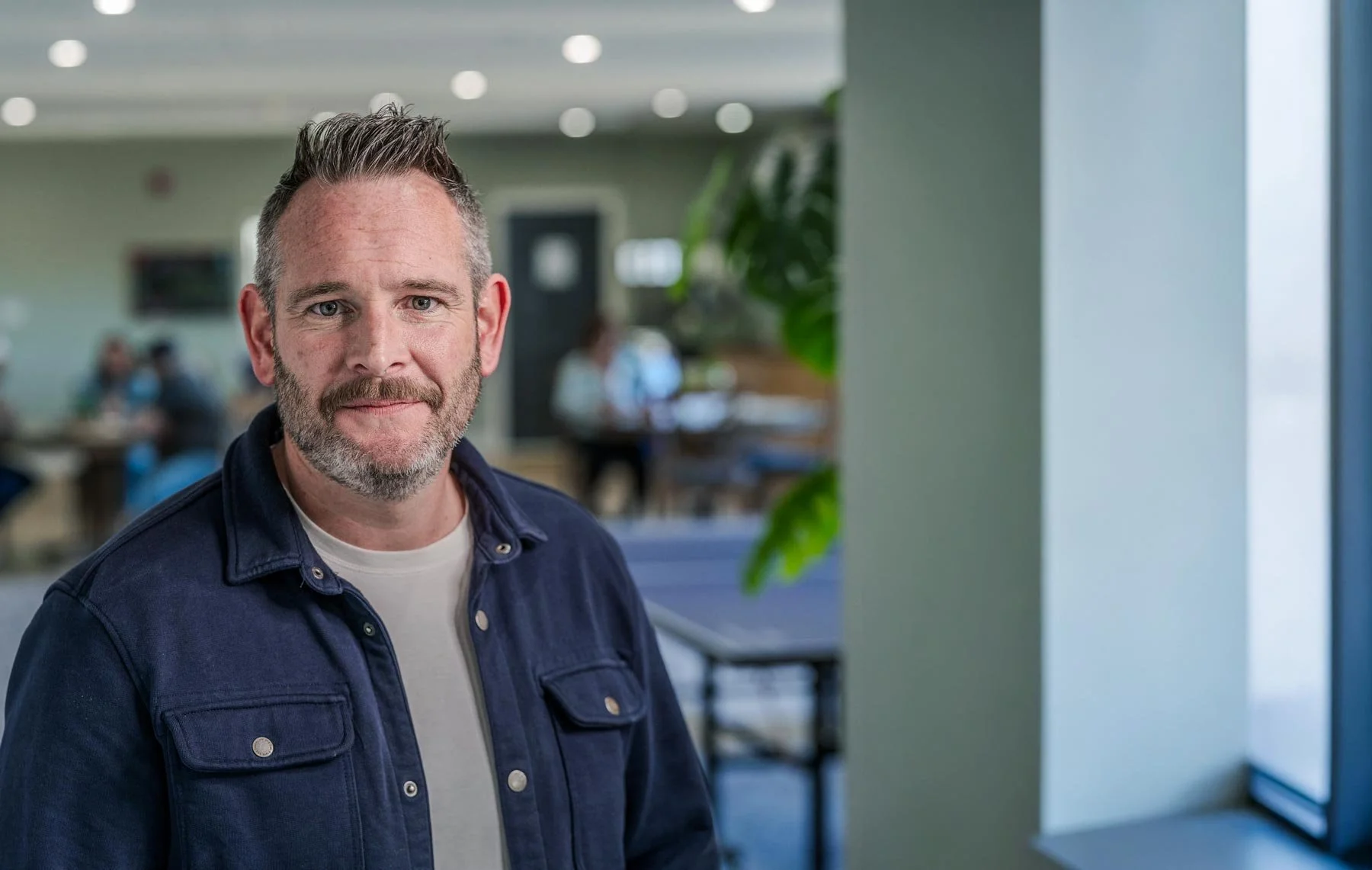 A middle-aged man with short, gray hair and a beard, wearing a navy blue jacket over a white shirt, standing in an office setting with people working at desks in the background, near a window with blinds.