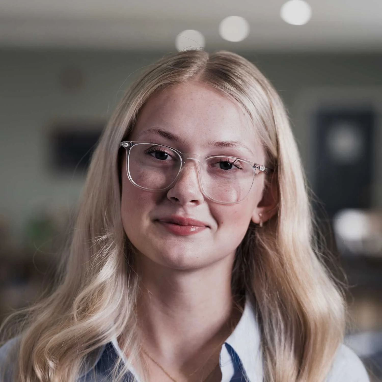A young woman with blonde hair, wearing clear glasses and a white collar shirt, in an indoor setting.