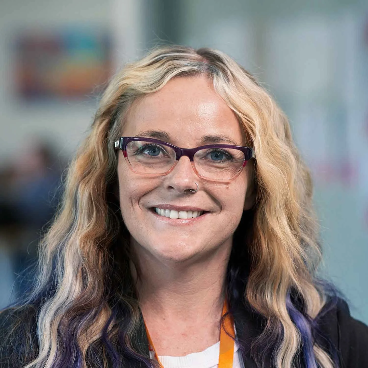Close-up of a woman with wavy blonde hair, purple glasses, and a bright smile, in an indoor setting.