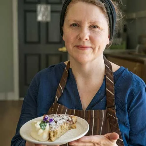 Woman in a blue shirt and striped apron holding a plate with a slice of dessert, whipped cream, and garnishes.