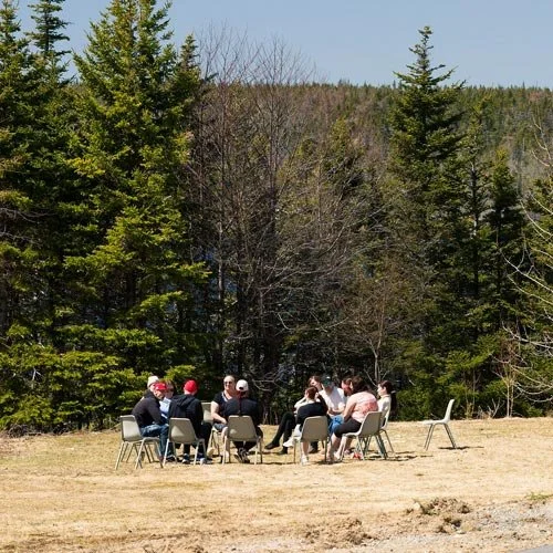 A group of people seated in a circle outdoors on a grassy field, at Vida Nova Recovery Centre. They are surrounded by trees with a forested hillside in the background.