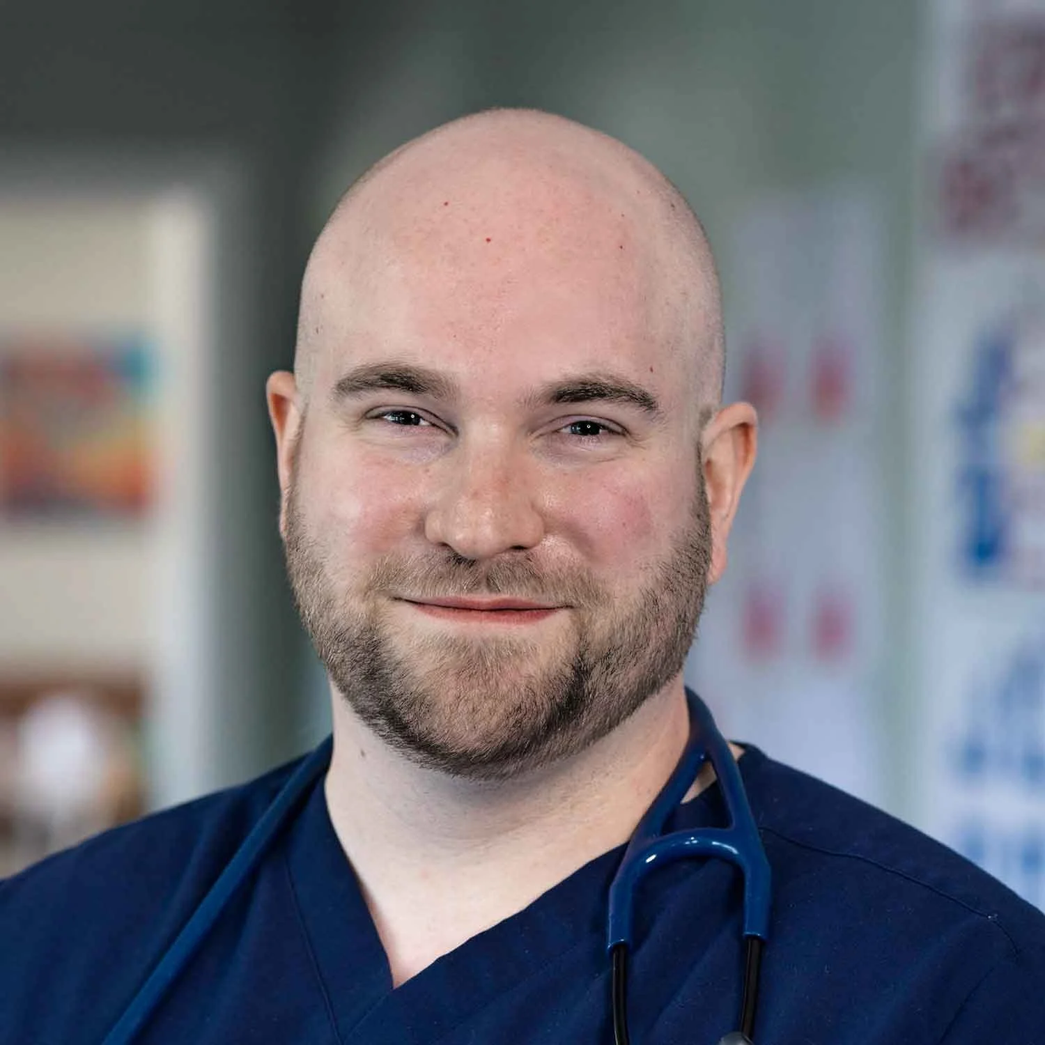 Portrait of a smiling man with a bald head and a beard, wearing dark medical scrubs and a stethoscope around his neck, in a medical setting.