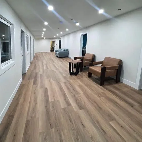 Long hallway with wood flooring, white walls, and recessed ceiling lights, furnished with a few armchairs and sofas.