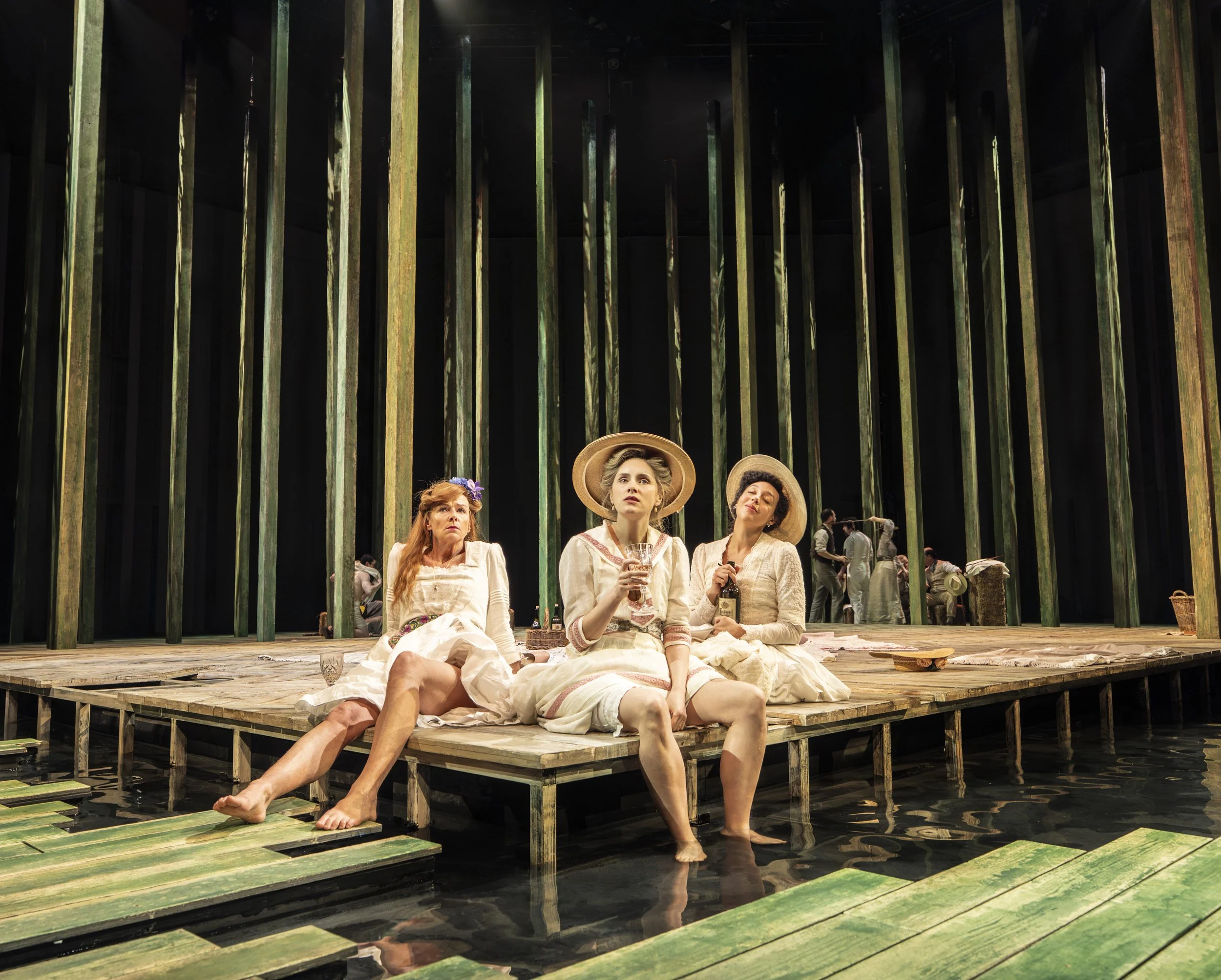 Three women dressed in vintage-style clothing sitting on a wooden platform over water, with water and green wooden pillars in the background. One woman is holding a glass, and they are wearing hats and floral accessories.