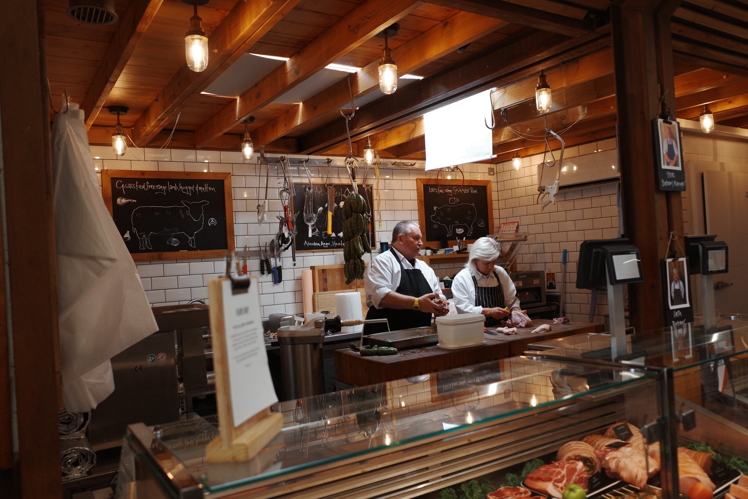 Inside a butcher shop, two workers are preparing meat behind the counter. There are blackboards with butcher diagrams and prices on the wall. Various cuts of meat and sausages are displayed on the counter and shelves. The shop has wooden accents and exposed lighting.