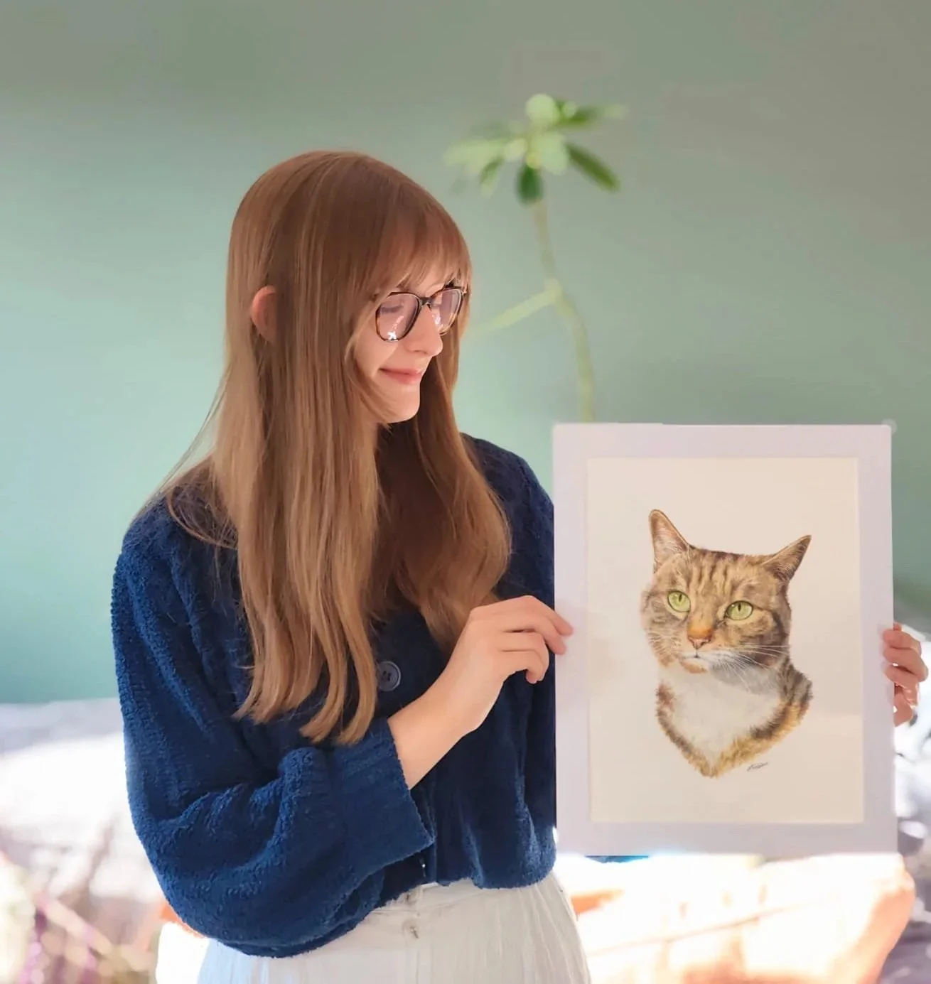 A woman with long, blonde hair and glasses is holding a framed picture of a tabby cat with green eyes.
