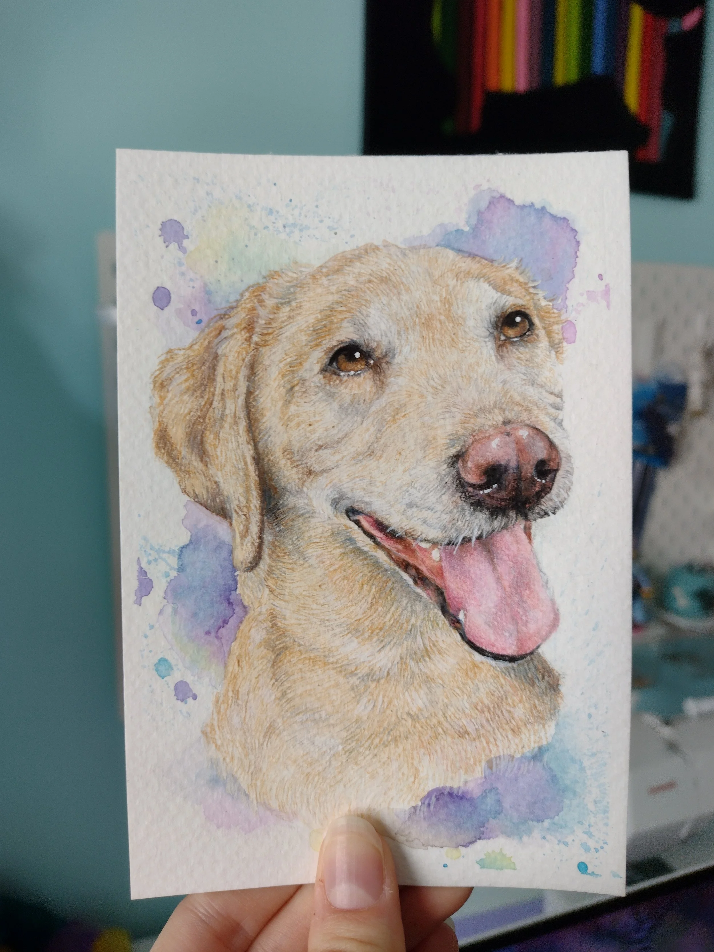 Hand holding a watercolor poster of a happy golden retriever dog with a pink tongue and expressive eyes, with a colorful abstract background.