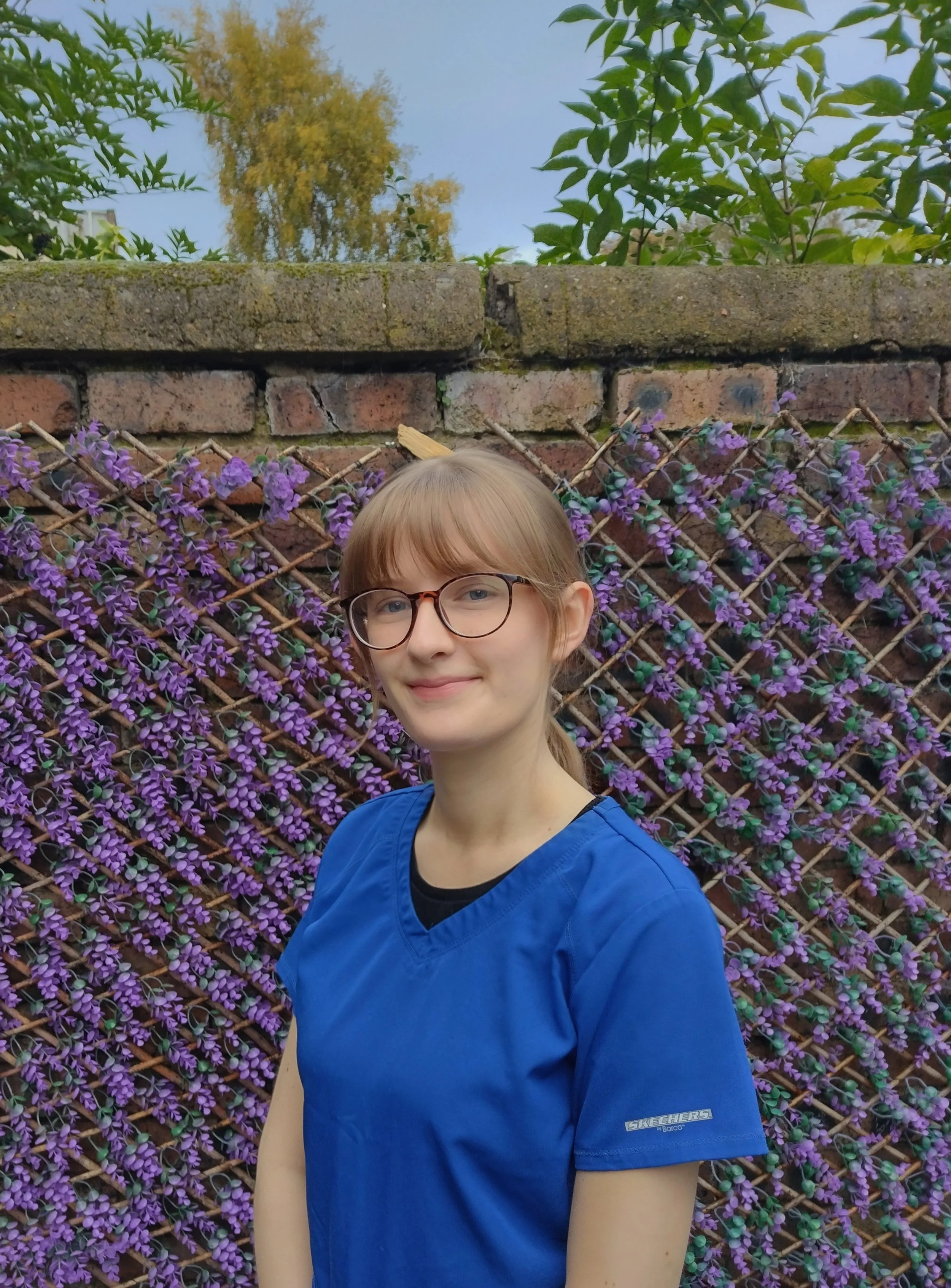 A woman with glasses and blue veterinary nursing scrubs is standing in front of a purple flower-covered fence and a brick wall, with trees and sky in the background.