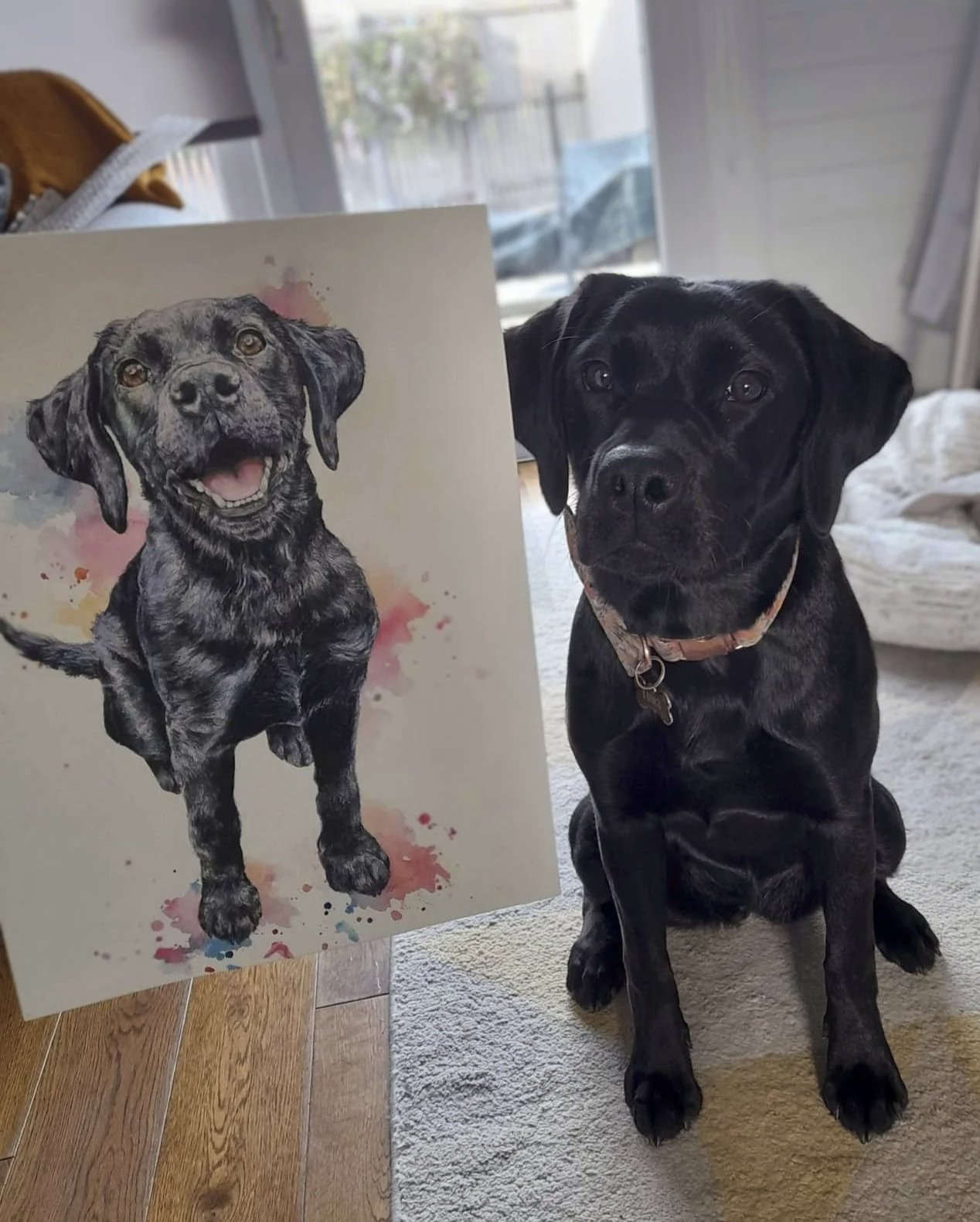 A black Labrador retriever puppy sitting on a beige carpet next to a watercolor-style painting of a happy, black and gray dog with floppy ears and a smiling expression.