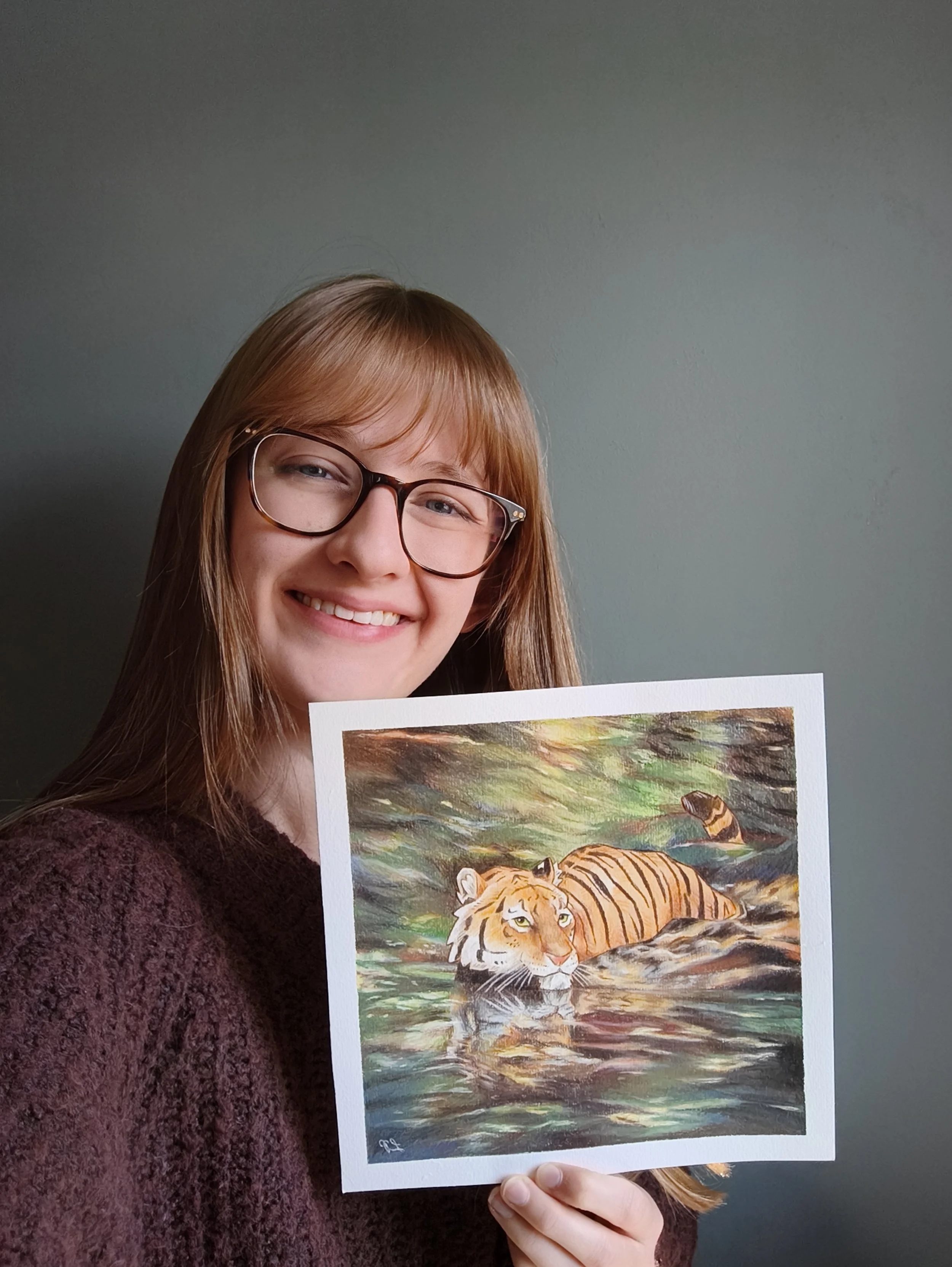 A person with blonde hair and glasses is smiling and holding a painting of a tiger in a river or pond with its reflection, surrounded by green and earthy tones.