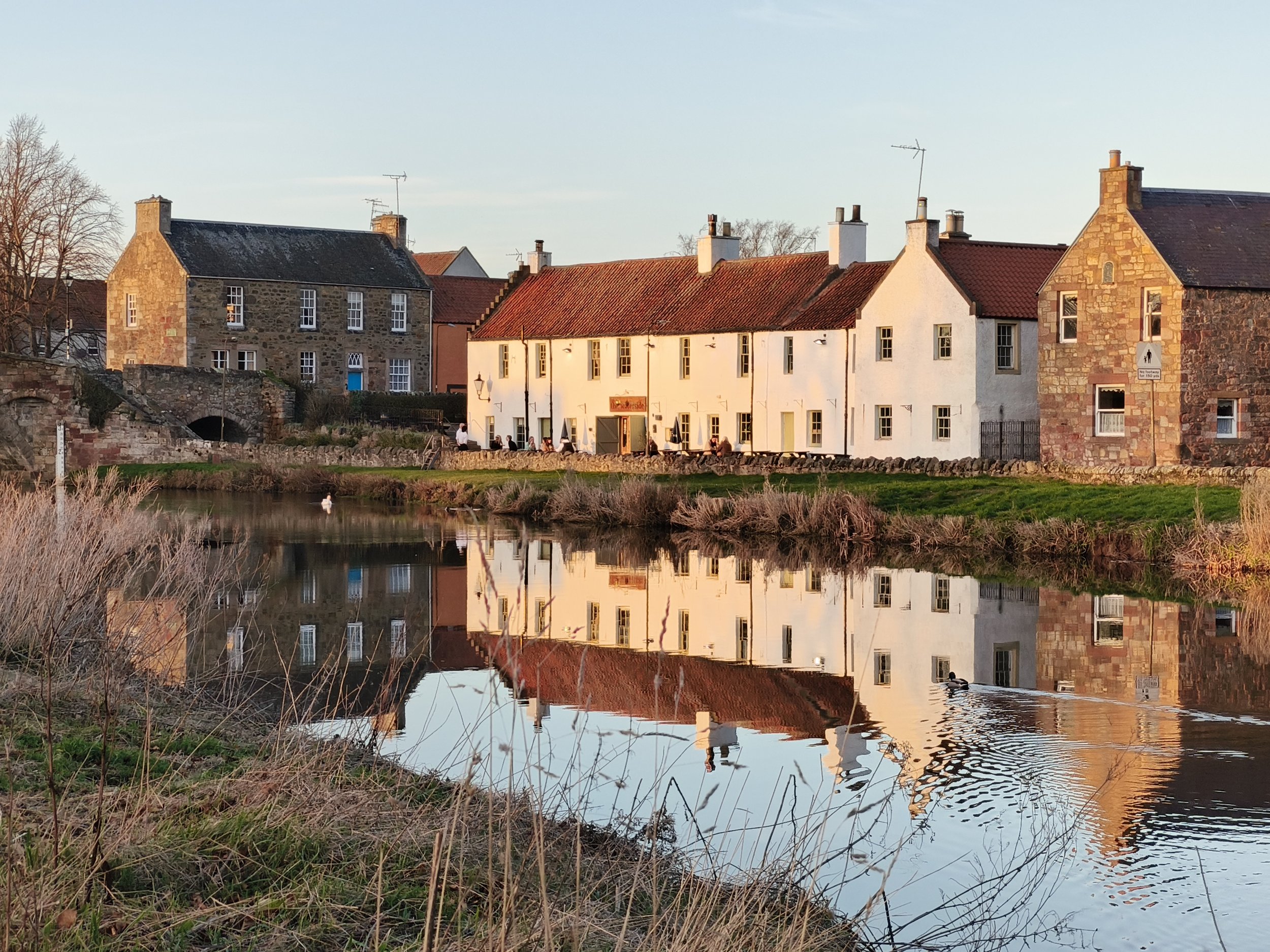 A row of houses with stone and white exteriors, red tile roofs, and chimneys, situated along a river with reflections. People are sitting outside at a cafe, and swans are swimming in the river during late afternoon or early evening.