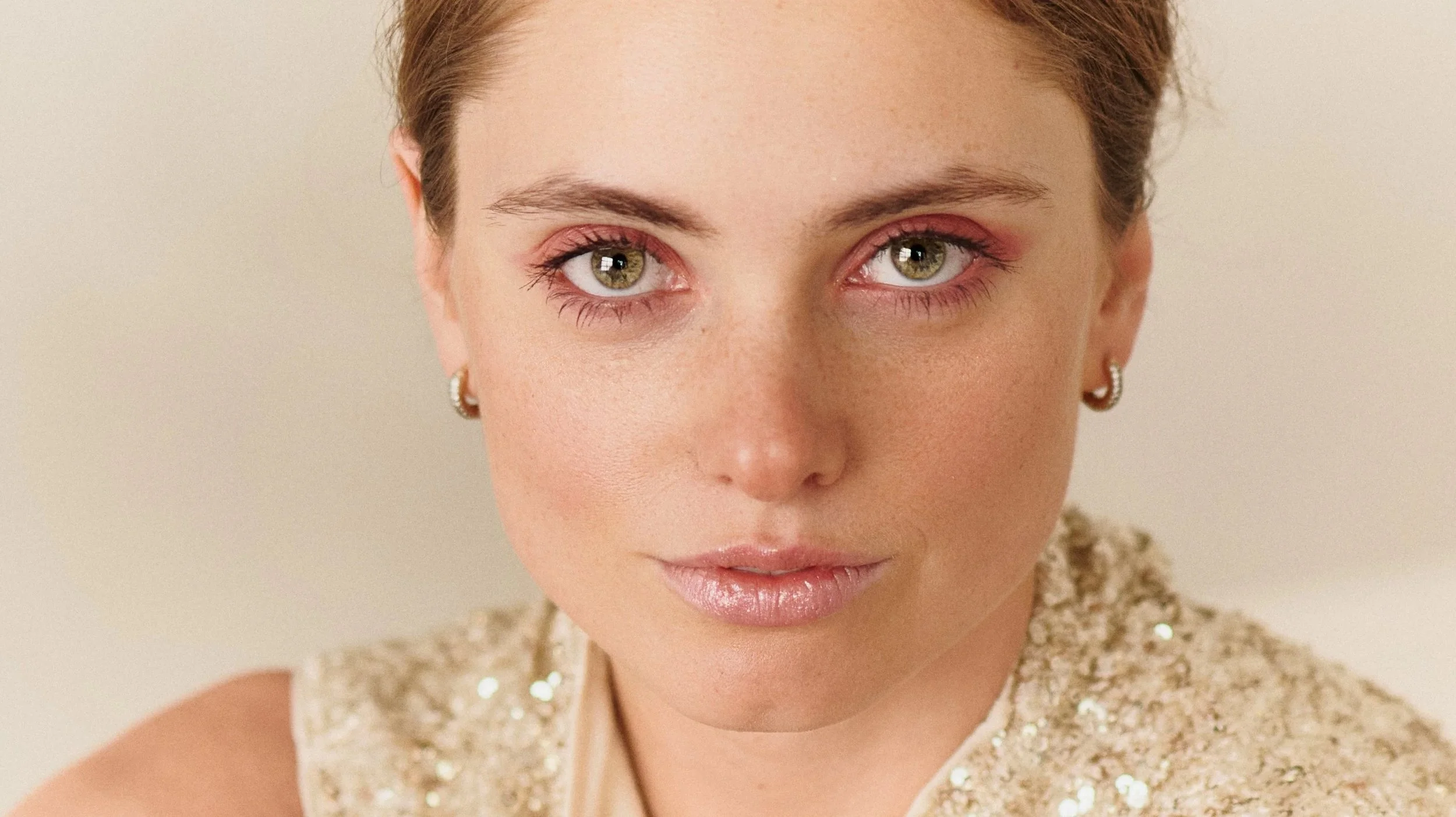 Close-up of a woman with short brown hair and green eyes wearing pink eye makeup and a glittering gold top, looking directly at the camera.