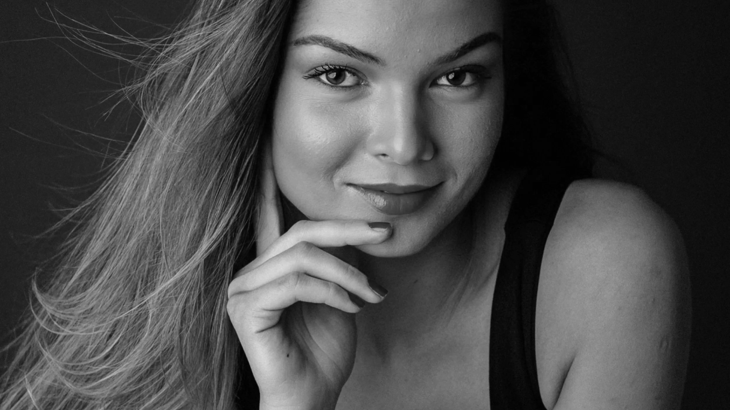 Close-up of a young woman with long hair touching her chin, looking at the camera, with a slight smile, in black and white.