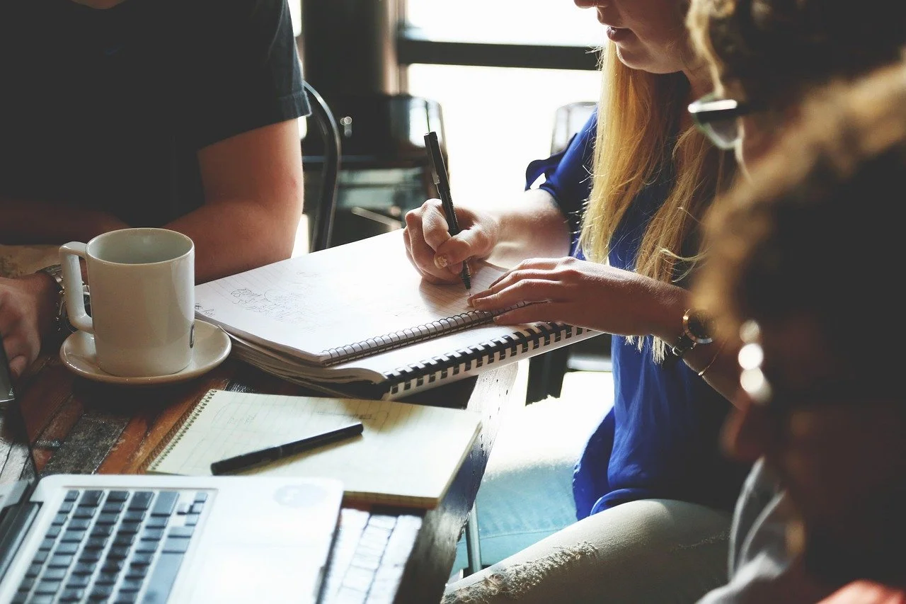 People working together at a table with notebooks, a pen, a cup of coffee, and a laptop.