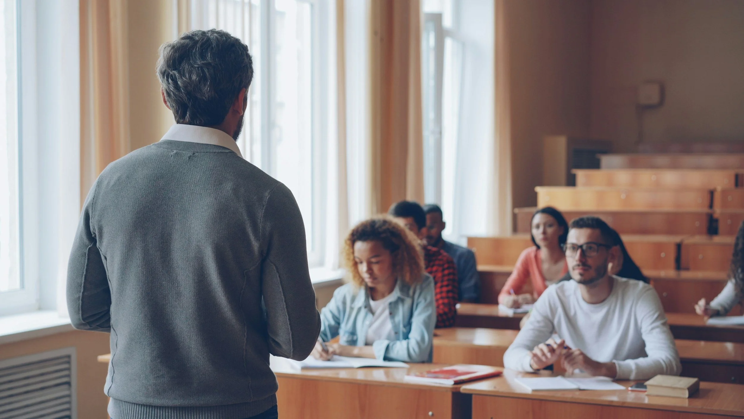 A man with dark hair wearing a gray sweater standing at the front of a classroom, facing a group of students seated at desks, with large windows and wooden tiered seating in the background.