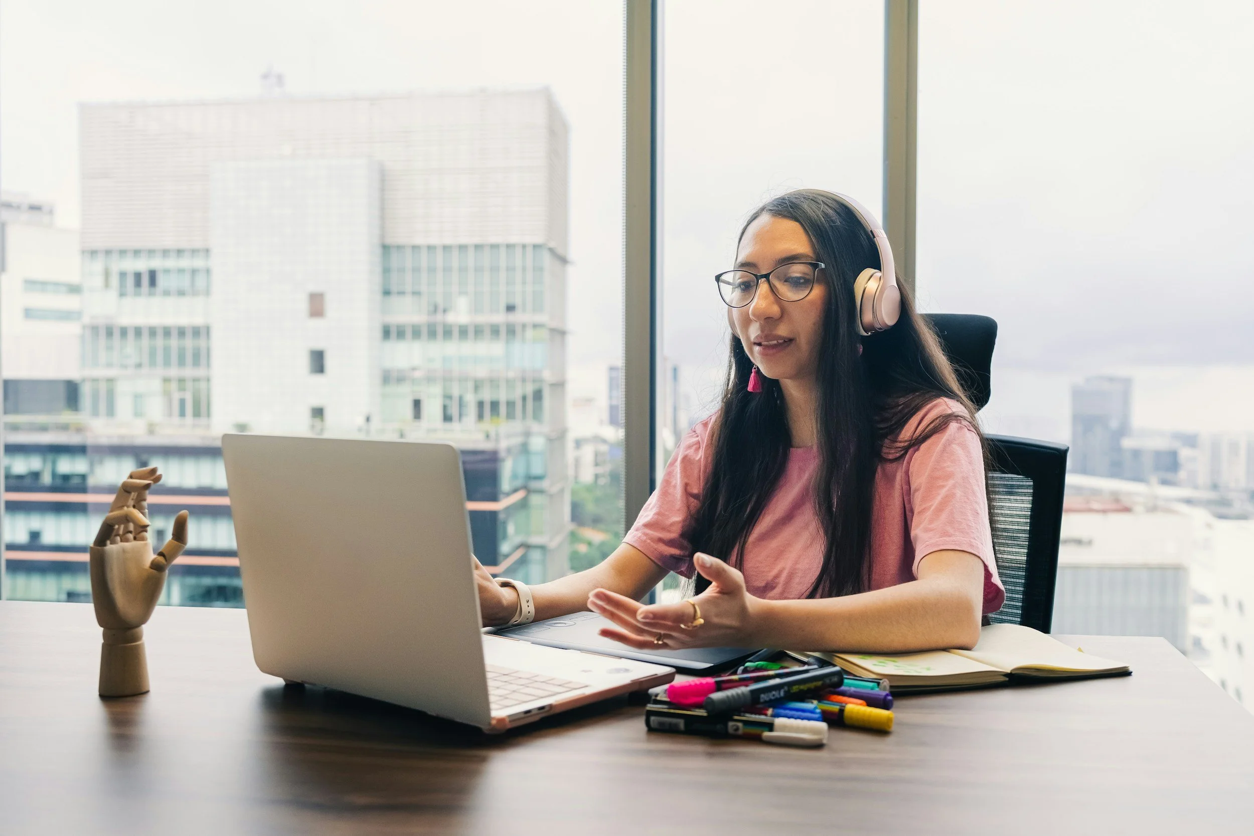 A young woman with long dark hair and glasses, wearing a pink shirt, sitting at a desk with a laptop, a notebook, and colorful markers, in an office with large windows overlooking city buildings.