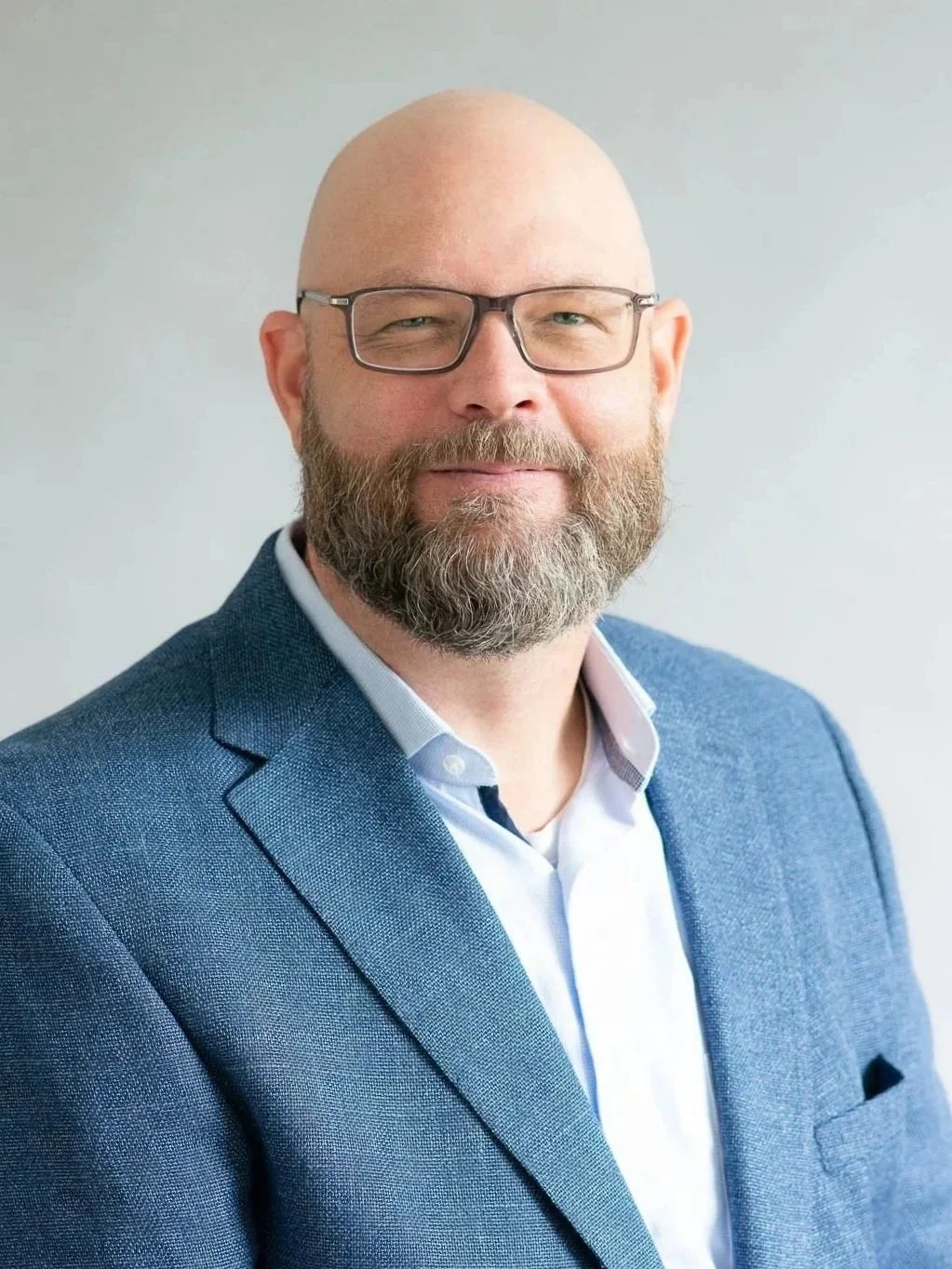 A professional man with glasses and a beard, wearing a blue blazer and white shirt, smiling against a plain background.