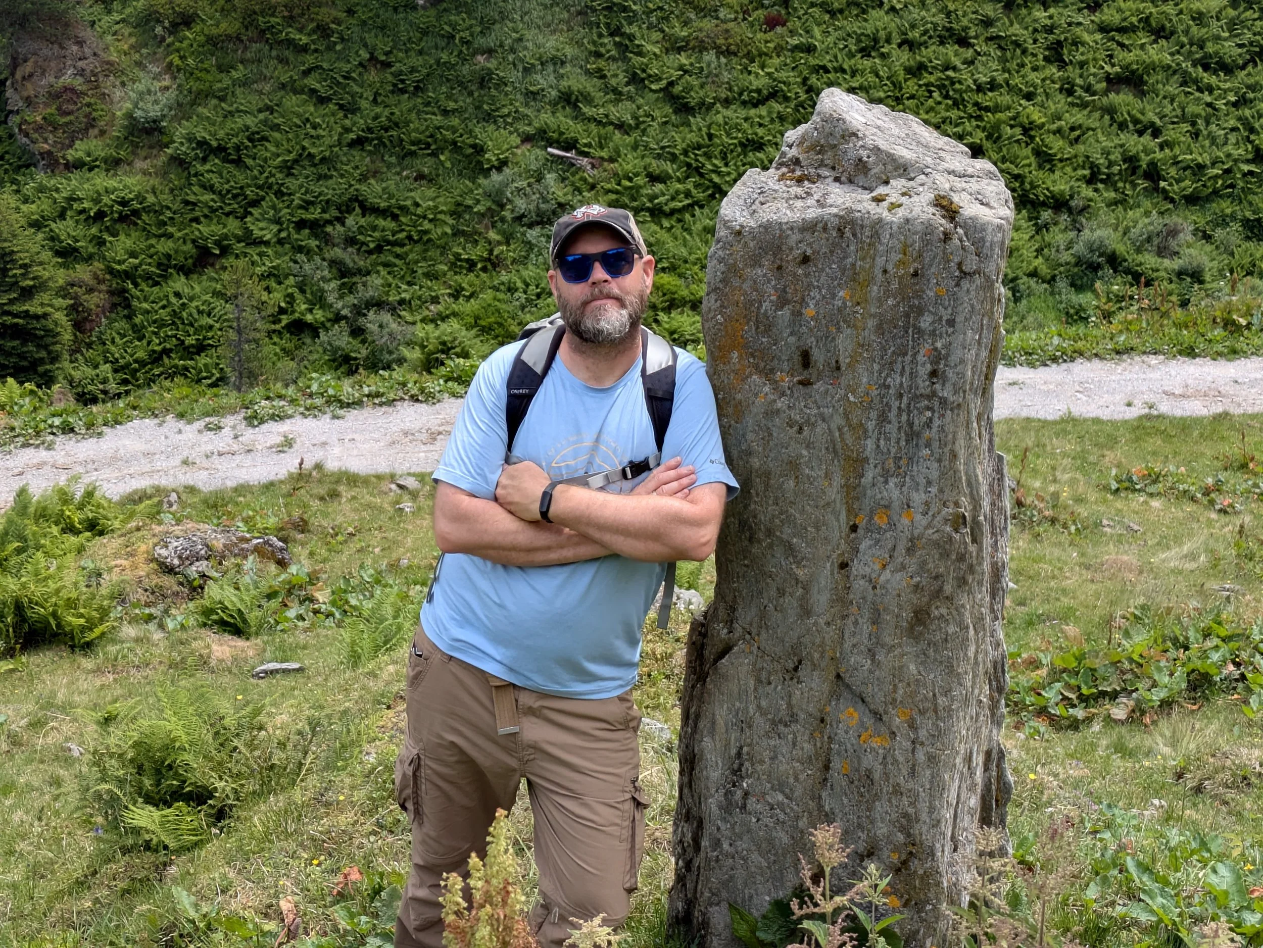 A man with sunglasses, a cap, and a backpack stands beside a large, moss-covered stone in a lush green landscape.