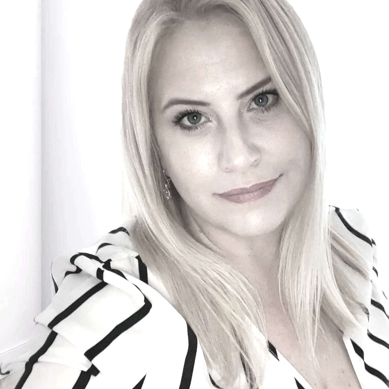 A woman with long blonde hair, wearing a white and black striped shirt, looking at the camera in front of a plain white background.