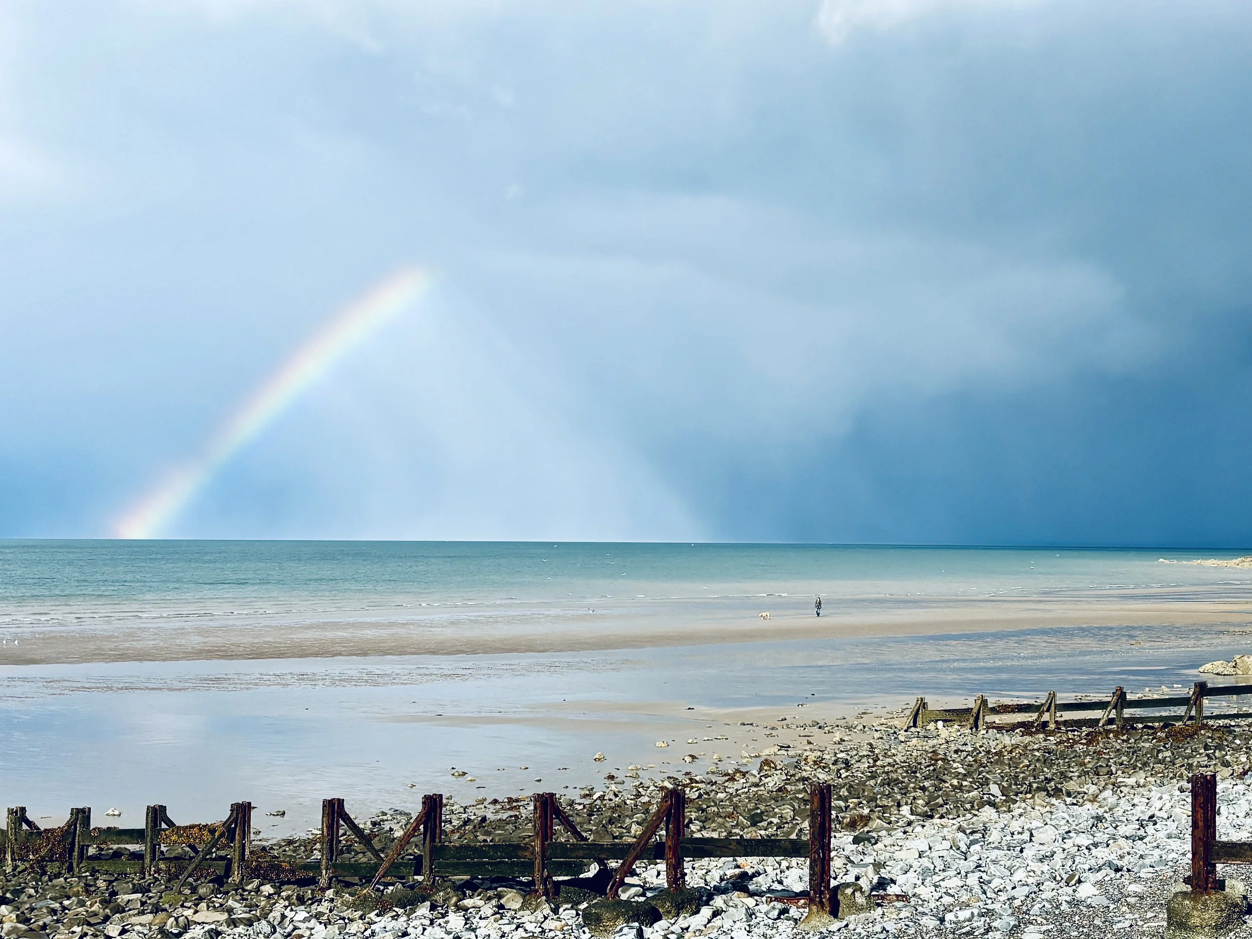 A beach scene with rocks along the shore, a wooden fence in the foreground, the ocean in the middle, and a cloudy sky with a rainbow in the sky.