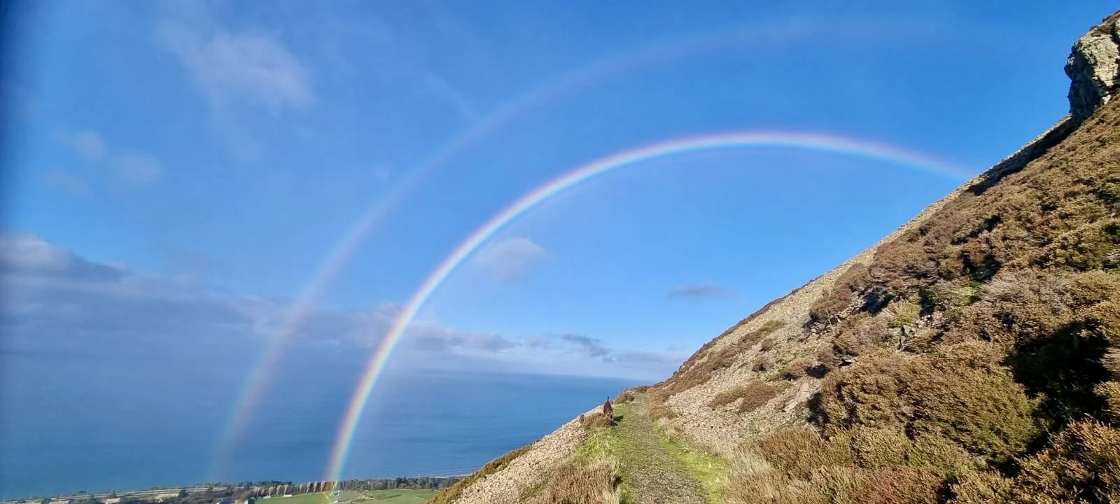 Double rainbow over a hillside with a path and coastal landscape in the distance.