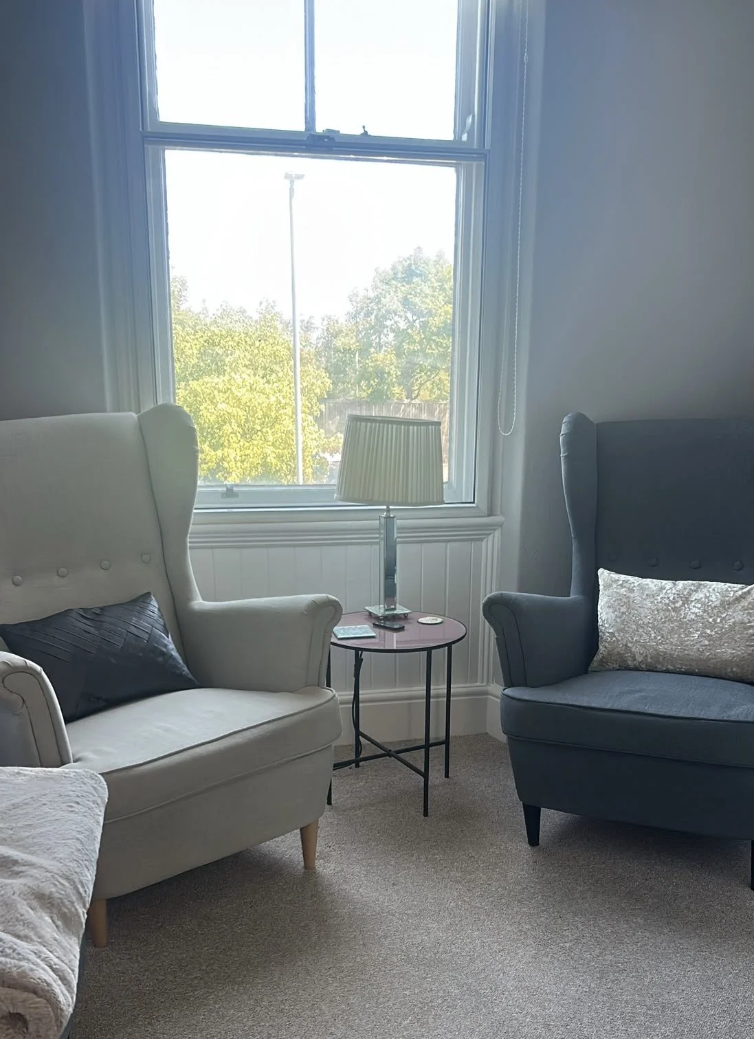 Living room with two armchairs, a side table with a lamp, and a window showing trees and a utility pole outside.