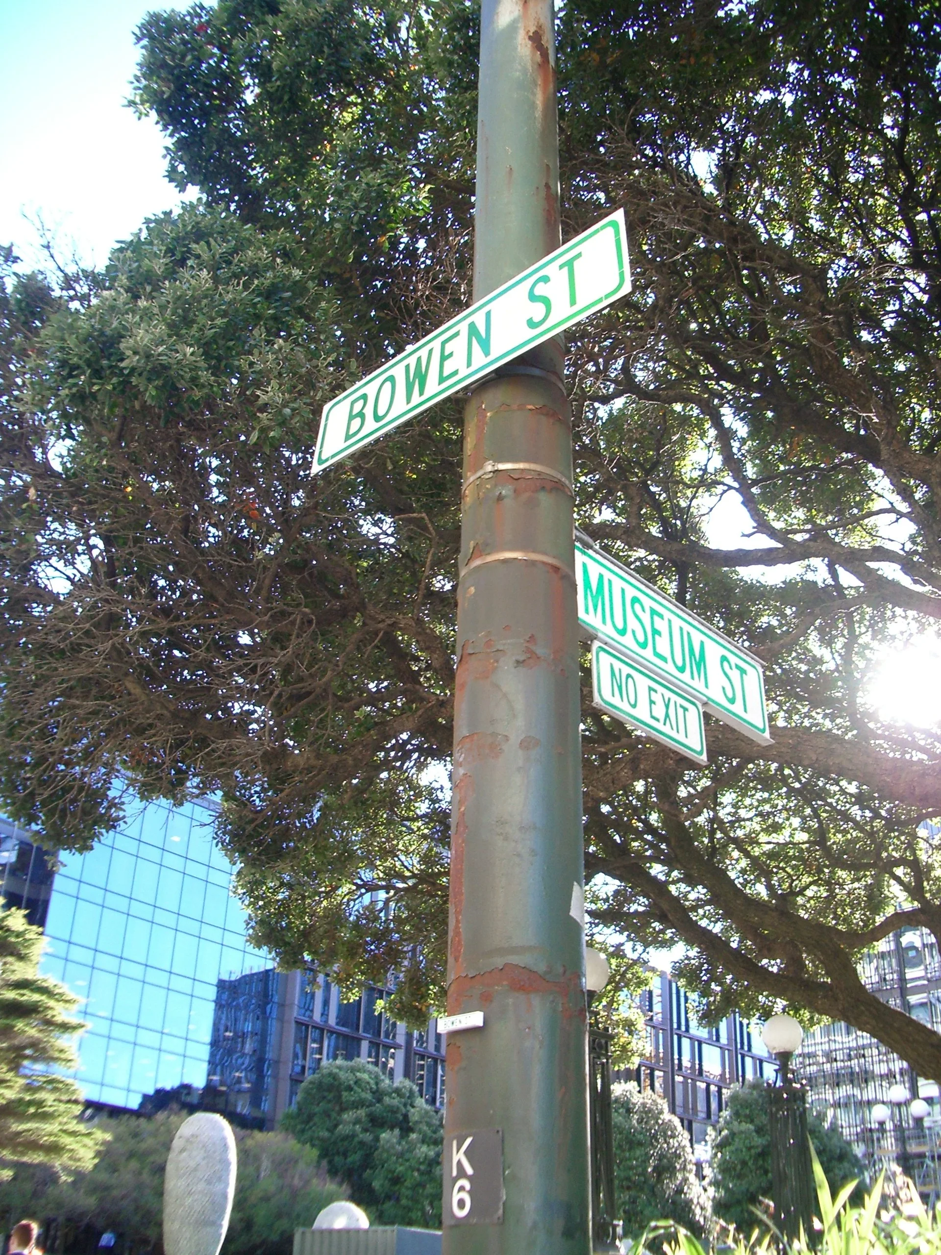 Street sign indicating 'BOWEN ST' and 'MUSEUM ST' with a 'NO EXIT' sign below, at an urban intersection with building glass windows, trees, and people in the background.