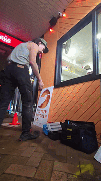 A man in a gray hoodie and black pants is cleaning the floor outside a fast food restaurant with a mop. There is an orange wooden wall, a window with a view inside, and a sign that reads 'BUTTER CHICKEN.' Various equipment and a yellow bottle are on the ground nearby.